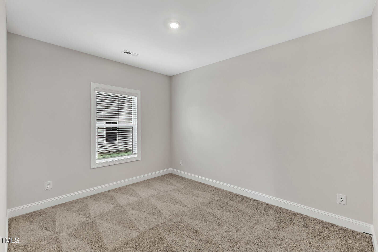 A small, empty room with light beige walls, a single window with white trim, tan carpeted flooring, and a ceiling light. Natural light comes through the window.
