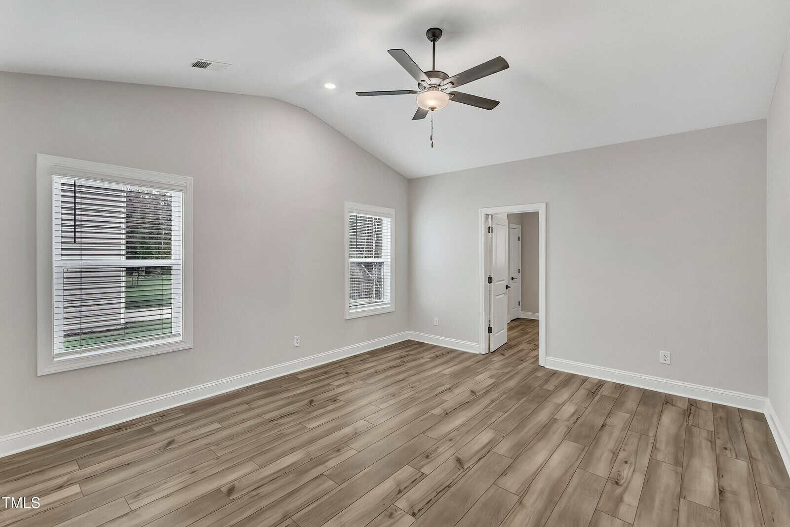 A bright, empty room with light gray walls, wood-style flooring, two windows with blinds, a ceiling fan with a light, and an open doorway leading to another room.