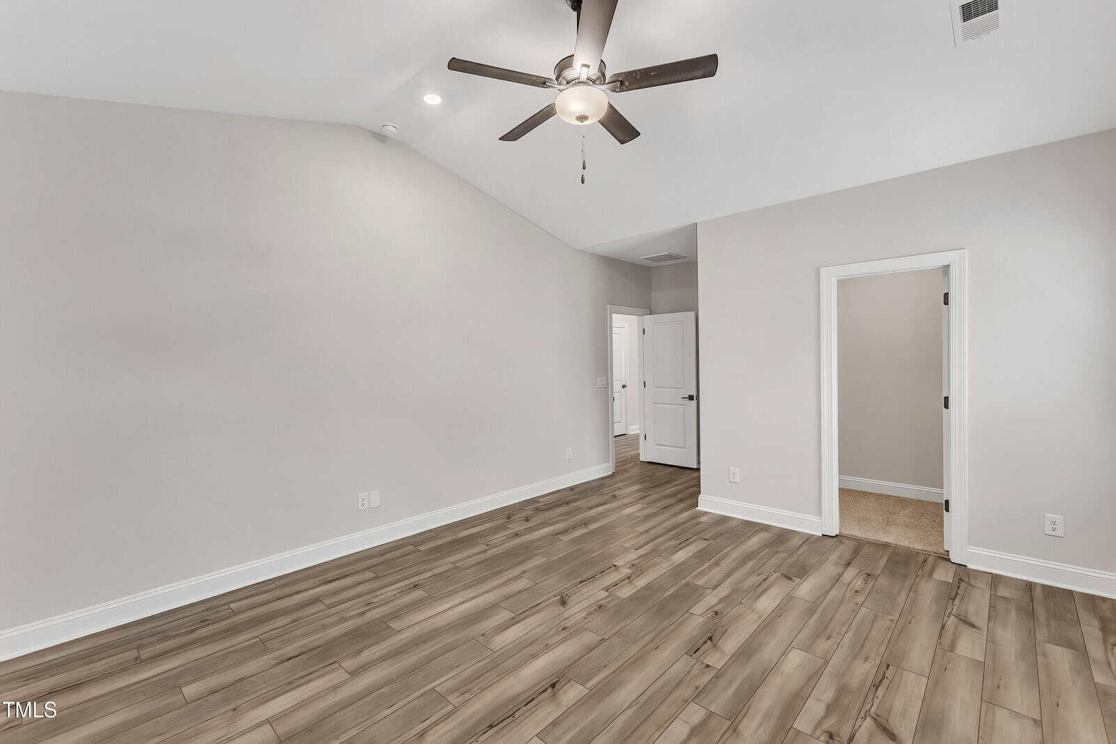 Spacious, empty room with light gray walls, wood-style flooring, a ceiling fan with lights, and a white door leading to an adjacent carpeted room; recessed lighting is also visible.
