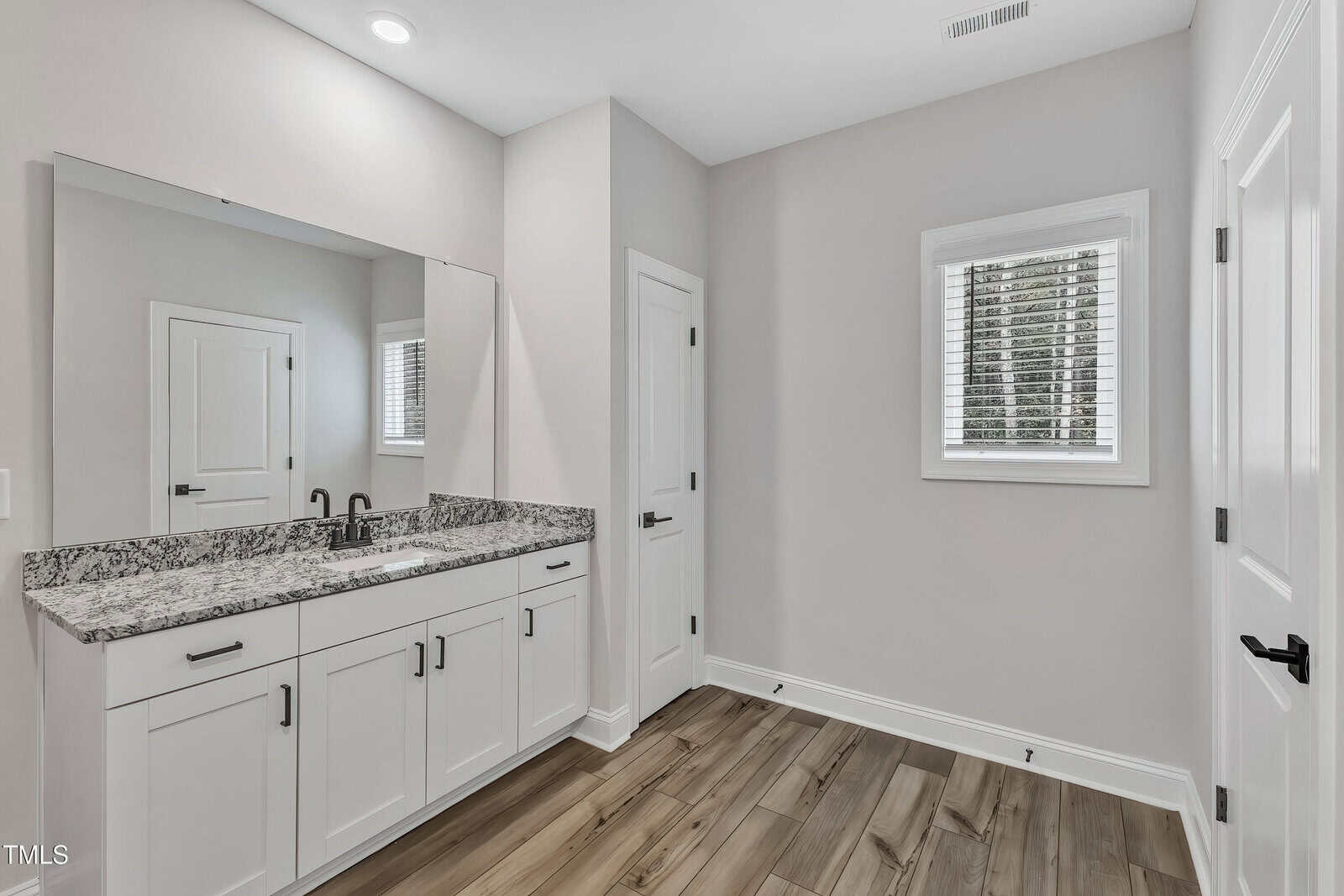 Modern bathroom with light gray walls, wood-look tile flooring, white vanity with granite countertop, single sink, large mirror, and a window with white blinds. Two white doors are also visible.