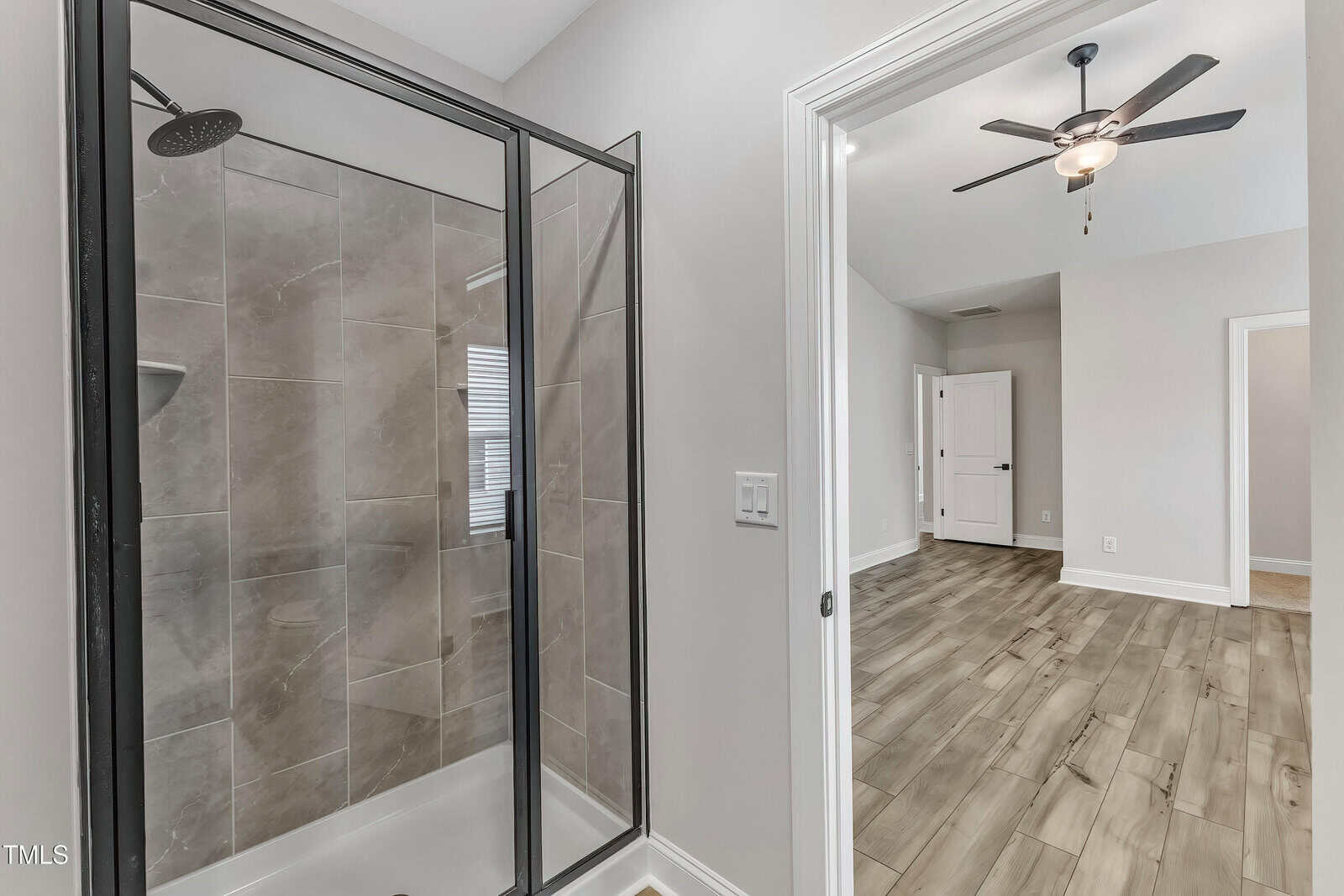 Modern bathroom with a glass shower enclosure and gray tile, next to a bedroom with wood flooring, white walls, a ceiling fan, and a white wardrobe.
