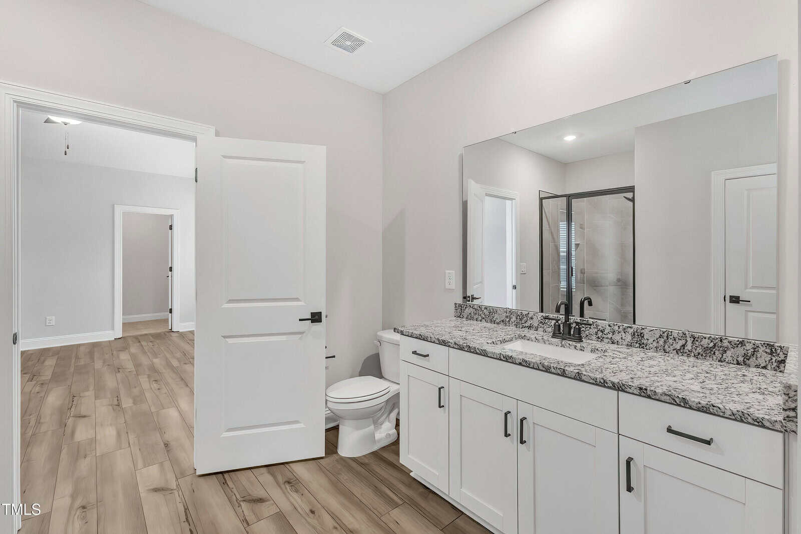 A modern bathroom with white cabinets, granite countertop, large mirror, and a toilet. The door is open, showing a wood-floored hallway and another room in the background.