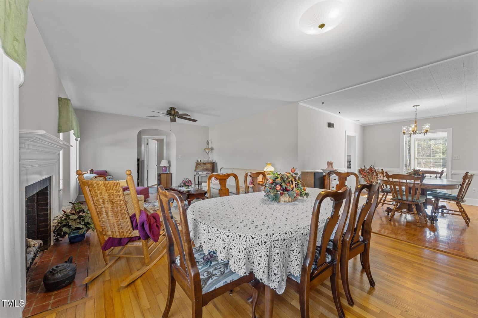 A spacious dining room with a wooden floor features a round table covered with a lace tablecloth and surrounded by chairs. In the background, there is another dining table and a fireplace. Various decorations and a ceiling fan are also visible.