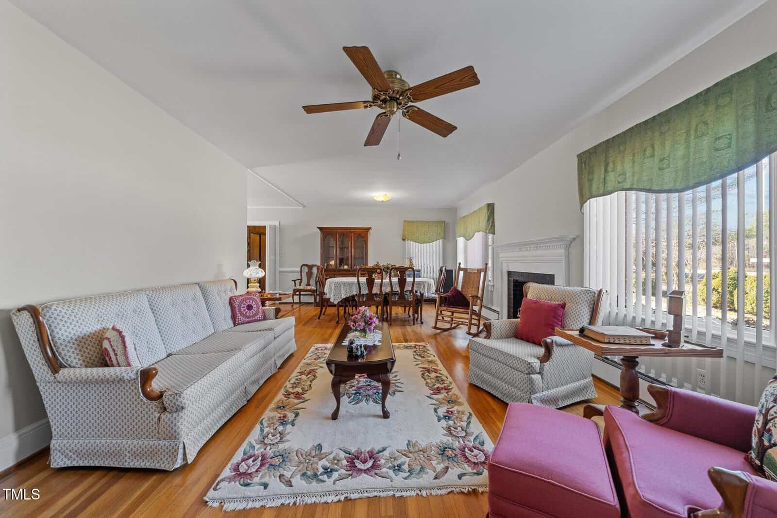 A living room with a floral rug, sofa, and two cushioned chairs. There's a wooden coffee table with flowers, a ceiling fan, and a dining table set in the background. Large windows provide natural light and green drapes adorn the windows.