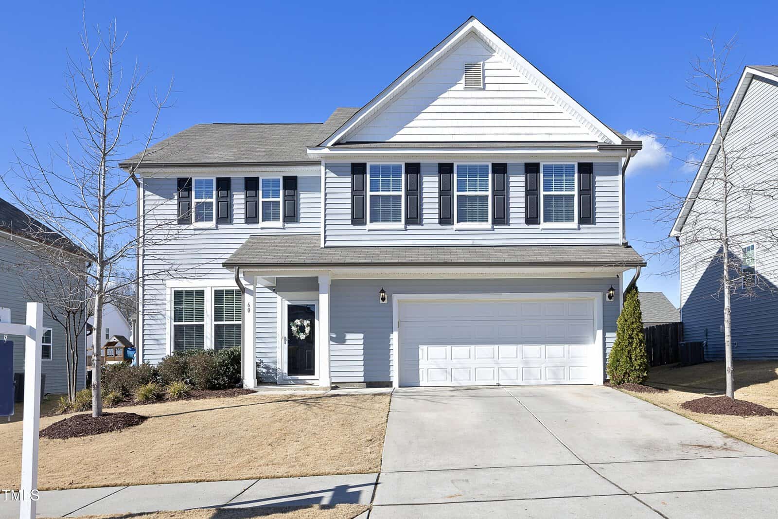 Two-story light gray house with white trim and black shutters, double garage, and a concrete driveway. The lawn is dry, and there are small leafless trees and shrubs in front of the house under a clear blue sky.