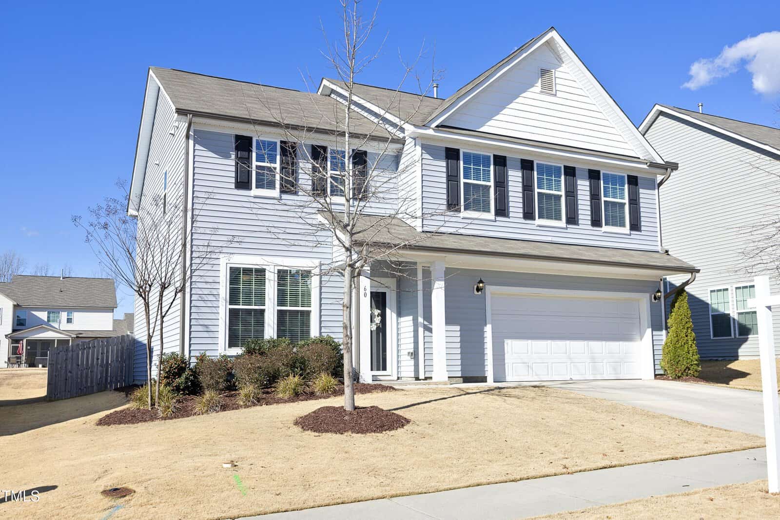 Two-story suburban house with light gray siding, black shutters, and a two-car garage. The front yard has sparse landscaping with small trees and bushes, and the lawn is dry. The sky is clear and blue.