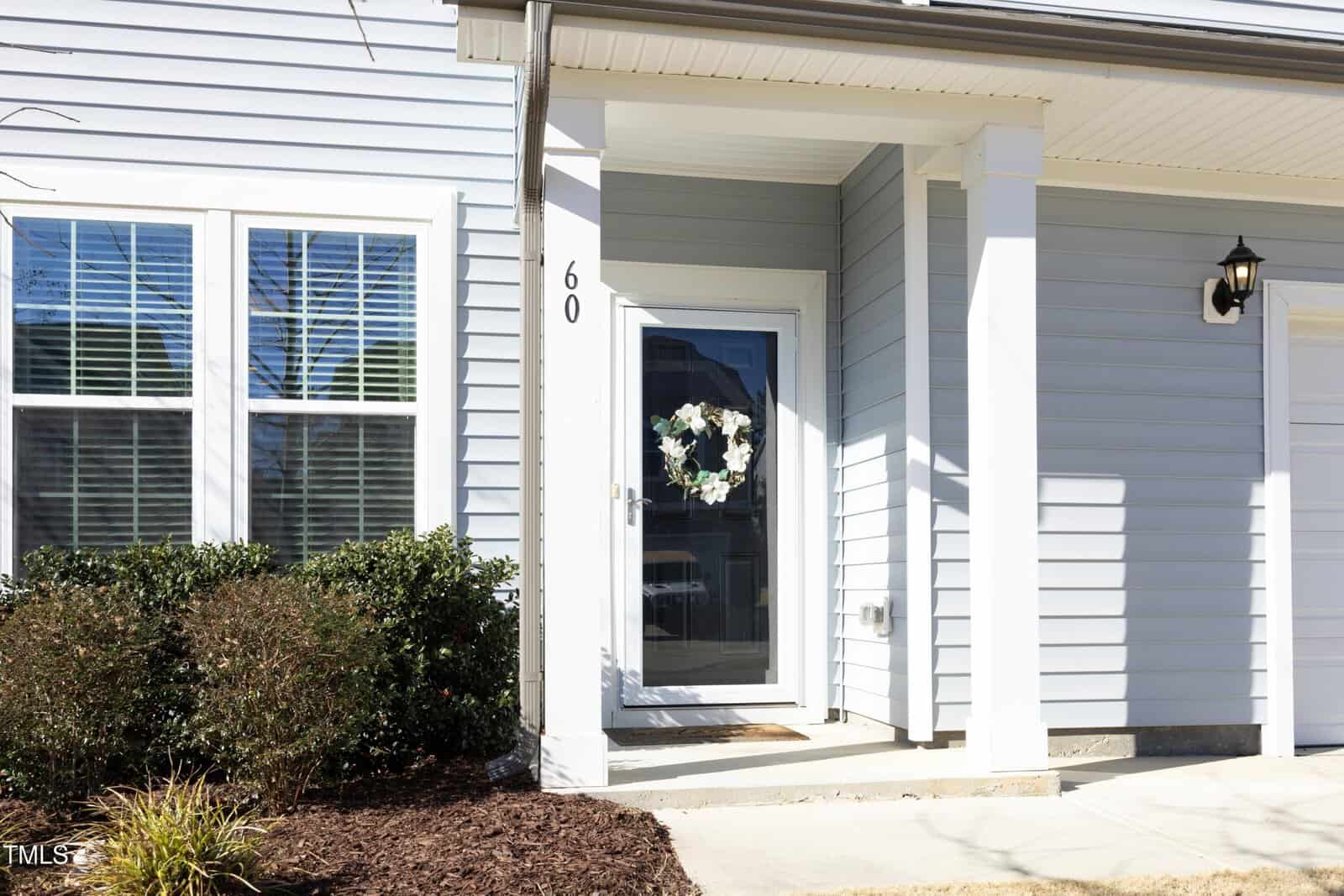 A light blue house with white trim featuring a front door adorned with a floral wreath, a white column, an exterior wall light, and two windows with closed blinds. Shrubs and mulch line the walkway to the entrance.