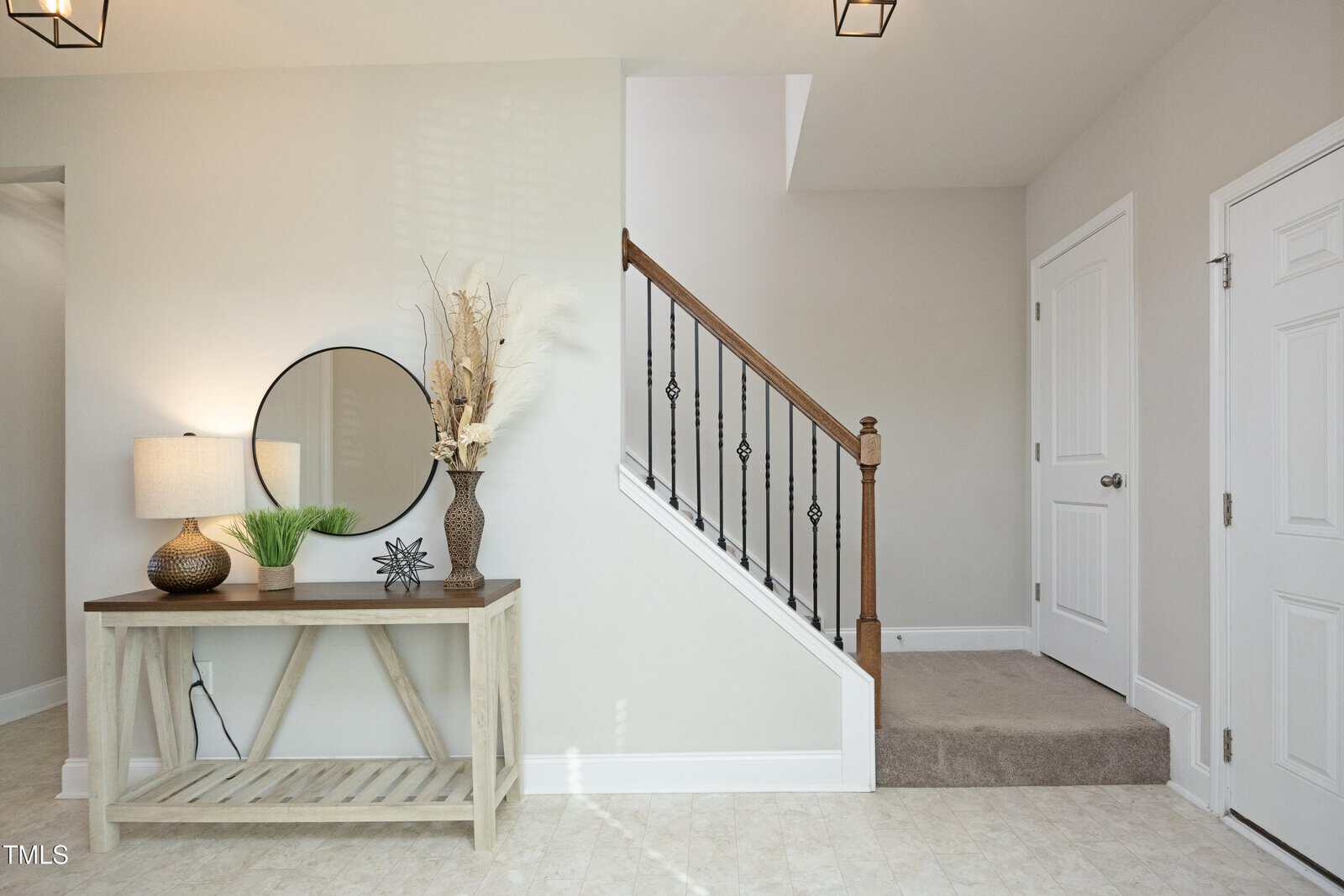 A small entryway with a light-colored console table holding a lamp, round mirror, decorative vase, and sculpture. A carpeted staircase with a wooden banister leads up, with two closed doors nearby.