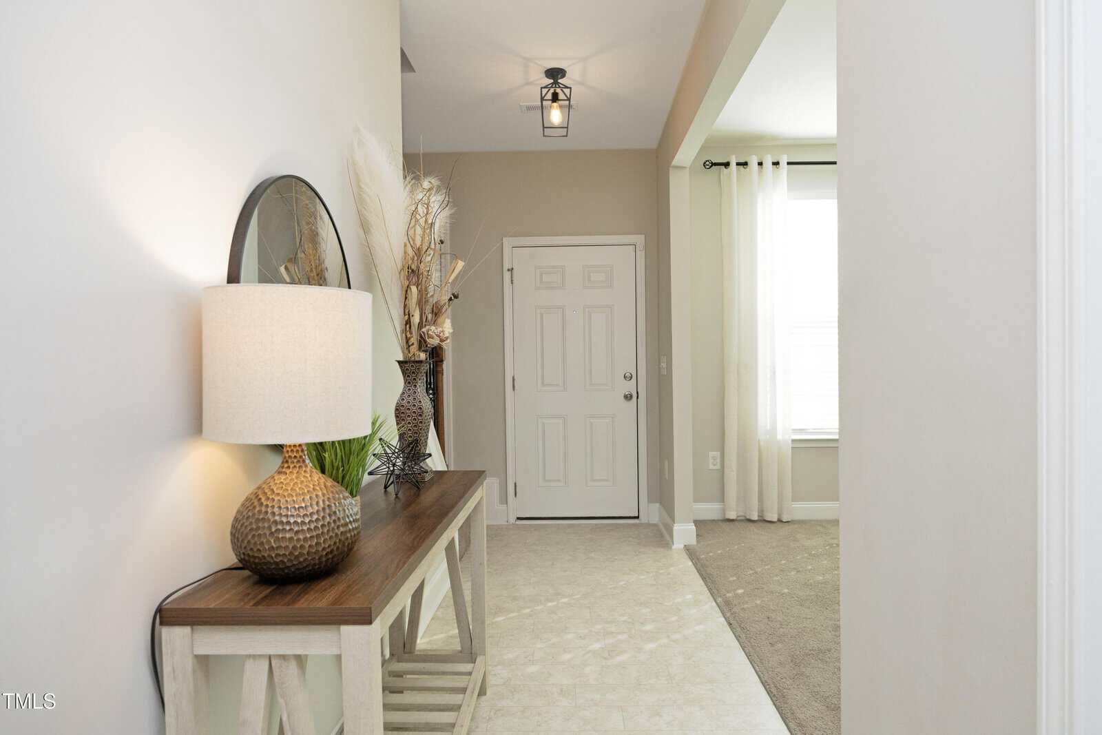 A hallway with a wooden console table holding a lamp, a round mirror, and decorative vases. A white front door is in the background, and a window with white curtains is on the right.