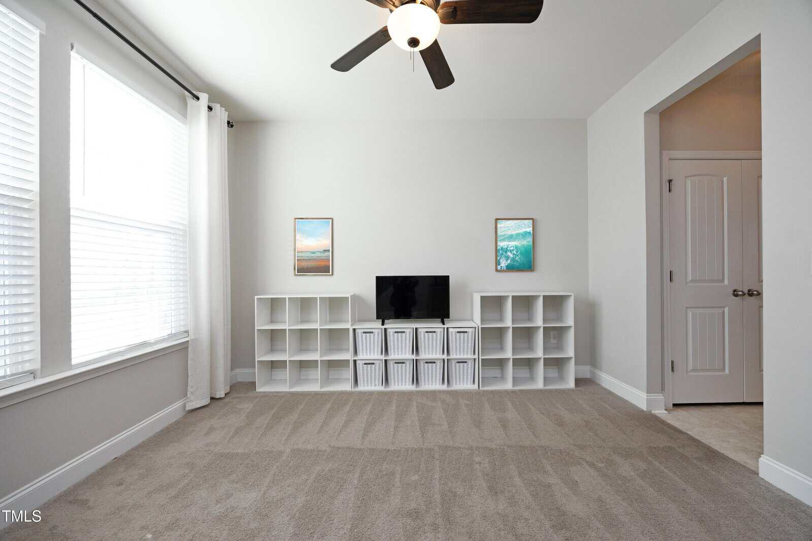 A bright room with beige carpet, white walls, a ceiling fan, a TV on white cube shelves, two framed ocean prints, and windows with white curtains on the left. A doorway and double doors are on the right.