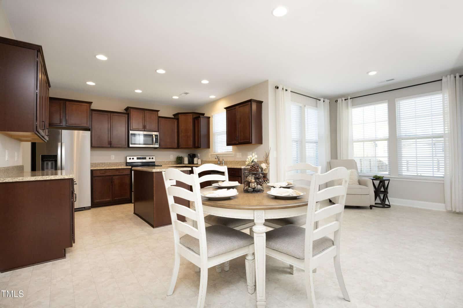 Bright kitchen with dark wood cabinets, stainless steel appliances, a central island, and a round dining table set for four. Large windows let in natural light, and a cozy armchair sits in the corner.