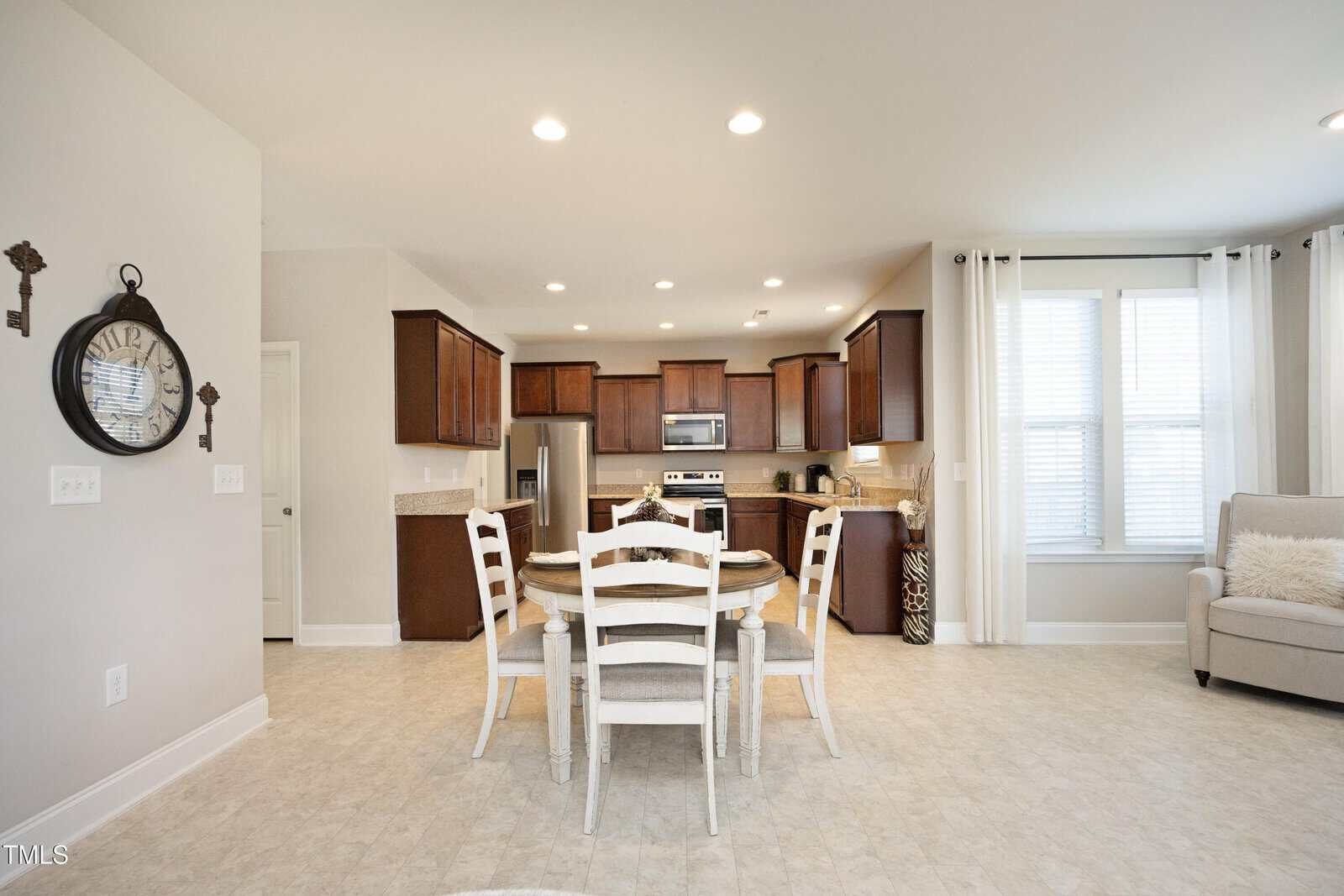 Bright kitchen and dining area with a round table and four white chairs, dark wood cabinets, stainless steel appliances, and a cozy armchair near large windows with white curtains.