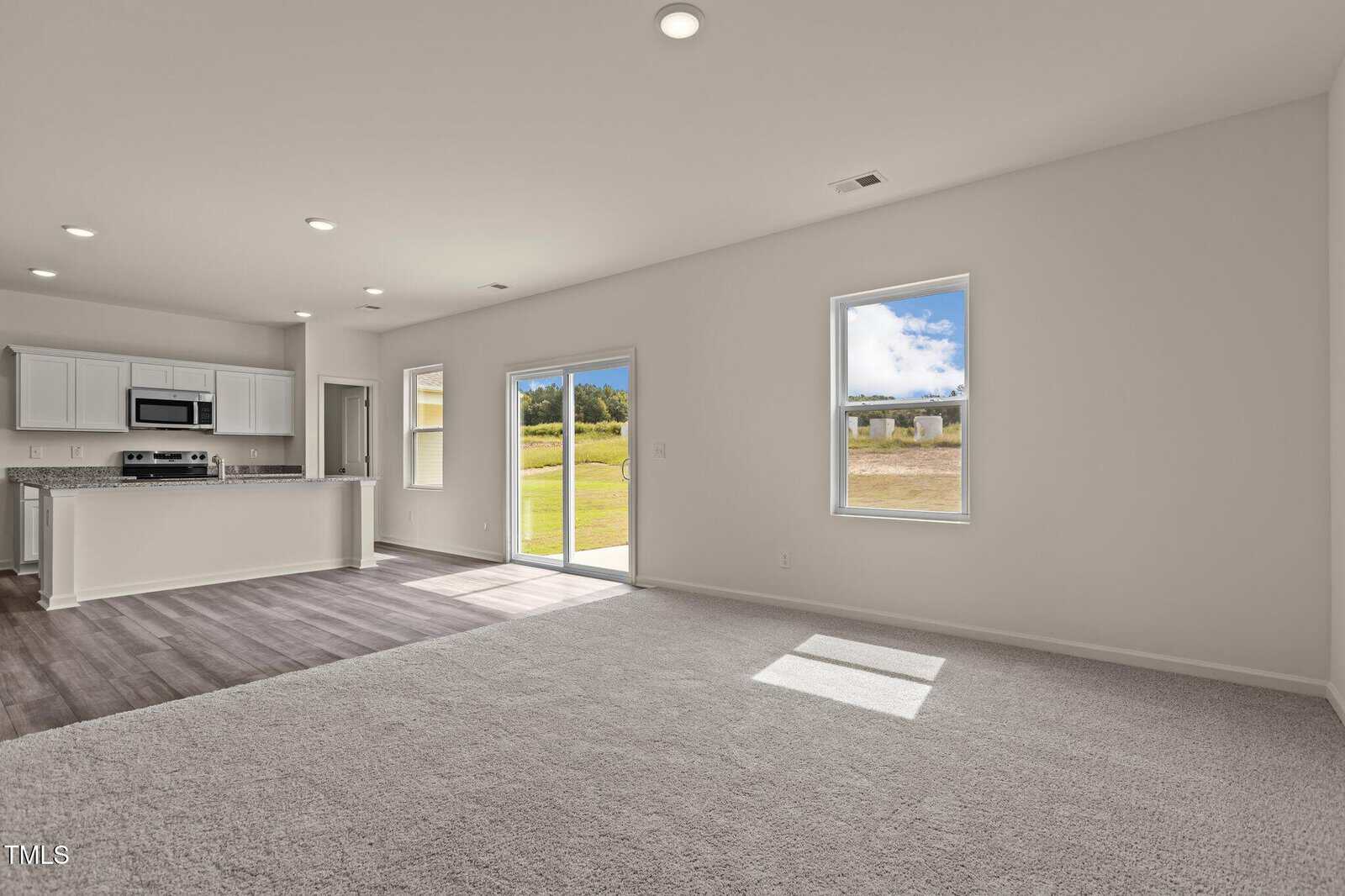 Spacious, unfurnished living room with carpeted floor, open to a modern kitchen with white cabinets and stainless steel appliances. Large windows and glass doors let in natural light with views of a grassy yard and blue sky.