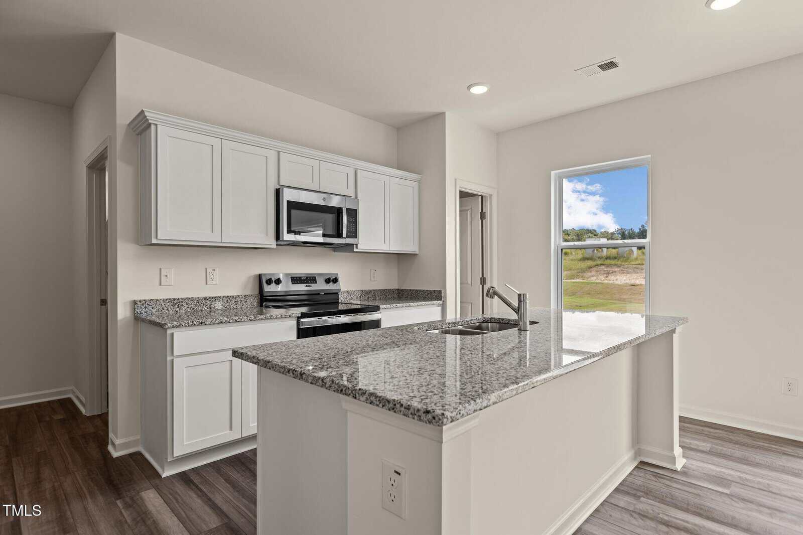 A modern kitchen with white cabinets, stainless steel appliances, granite countertops, wood flooring, and an island facing a window with a view of greenery outside.