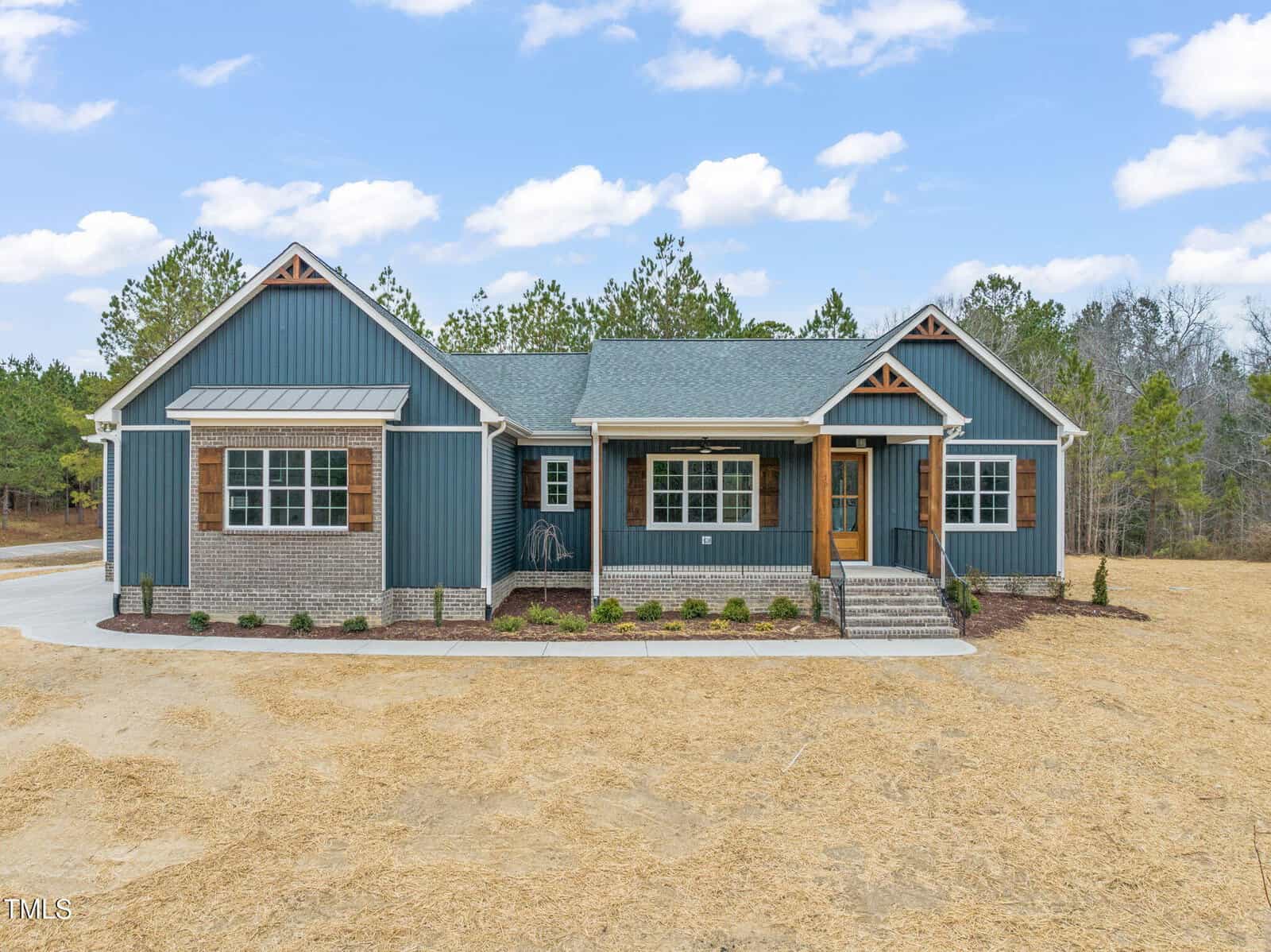 A modern single-story house with blue siding, brick accents, and wooden shutters. The home has a front porch with steps, surrounded by minimal landscaping, and sits on a large, bare lot with trees in the background.