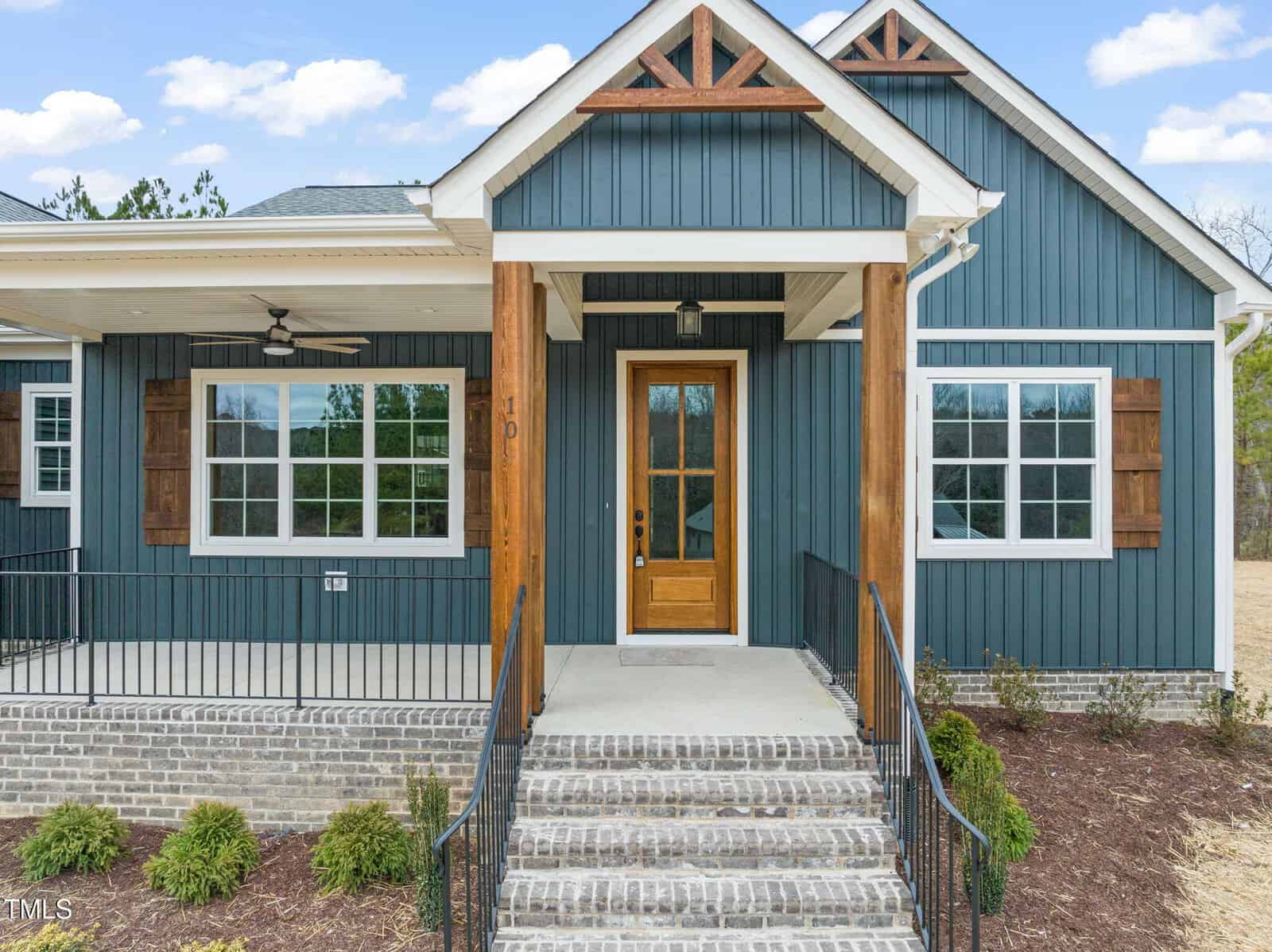 A modern blue house with white trim, wood accents, and a wooden front door. Brick steps lead to the entrance, flanked by metal railings and landscaped garden beds. Two large windows are on either side of the door.