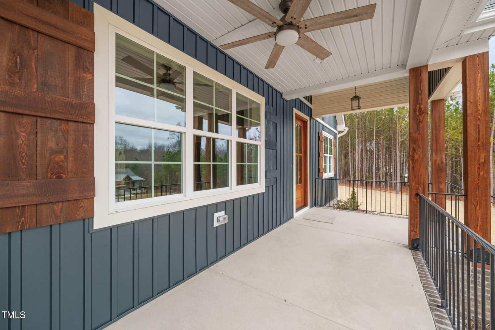 A spacious front porch with a ceiling fan and railing, featuring blue siding, brown wooden shutters, and a large window overlooking a wooded area.