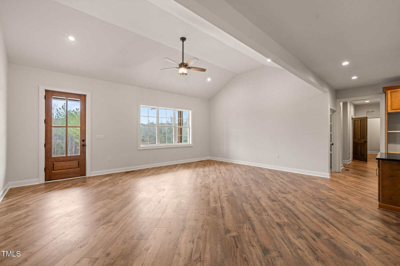 Spacious empty living room with wood flooring, large window, ceiling fan, and a wooden door leading outside. White walls and an open layout connect to a hallway and a kitchen area with visible wooden cabinetry.