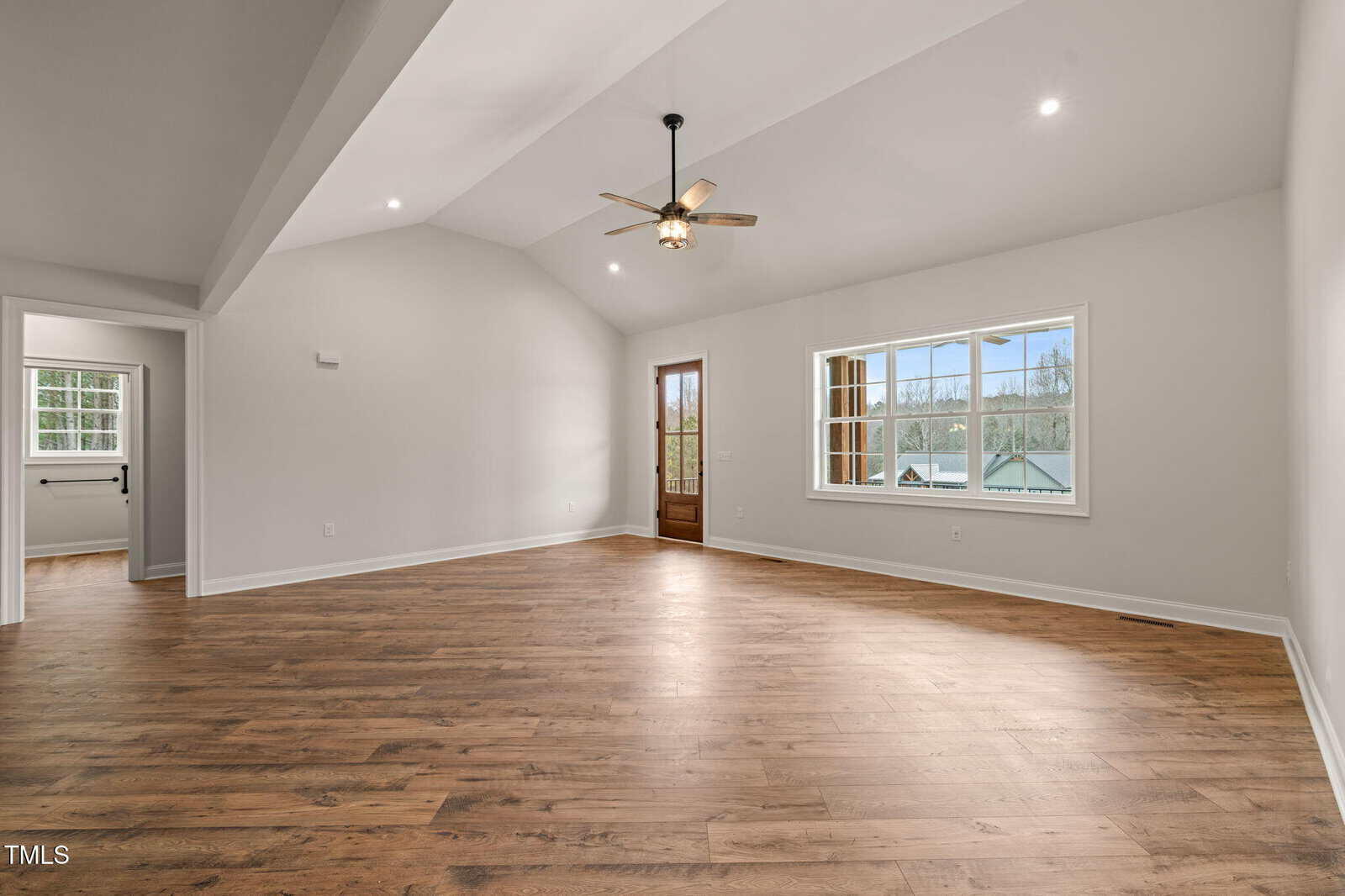 Spacious, empty living room with vaulted ceiling, wooden floor, beige walls, ceiling fan with light, large window letting in natural light, and a glass door to the outside. Doorway leads to another room.