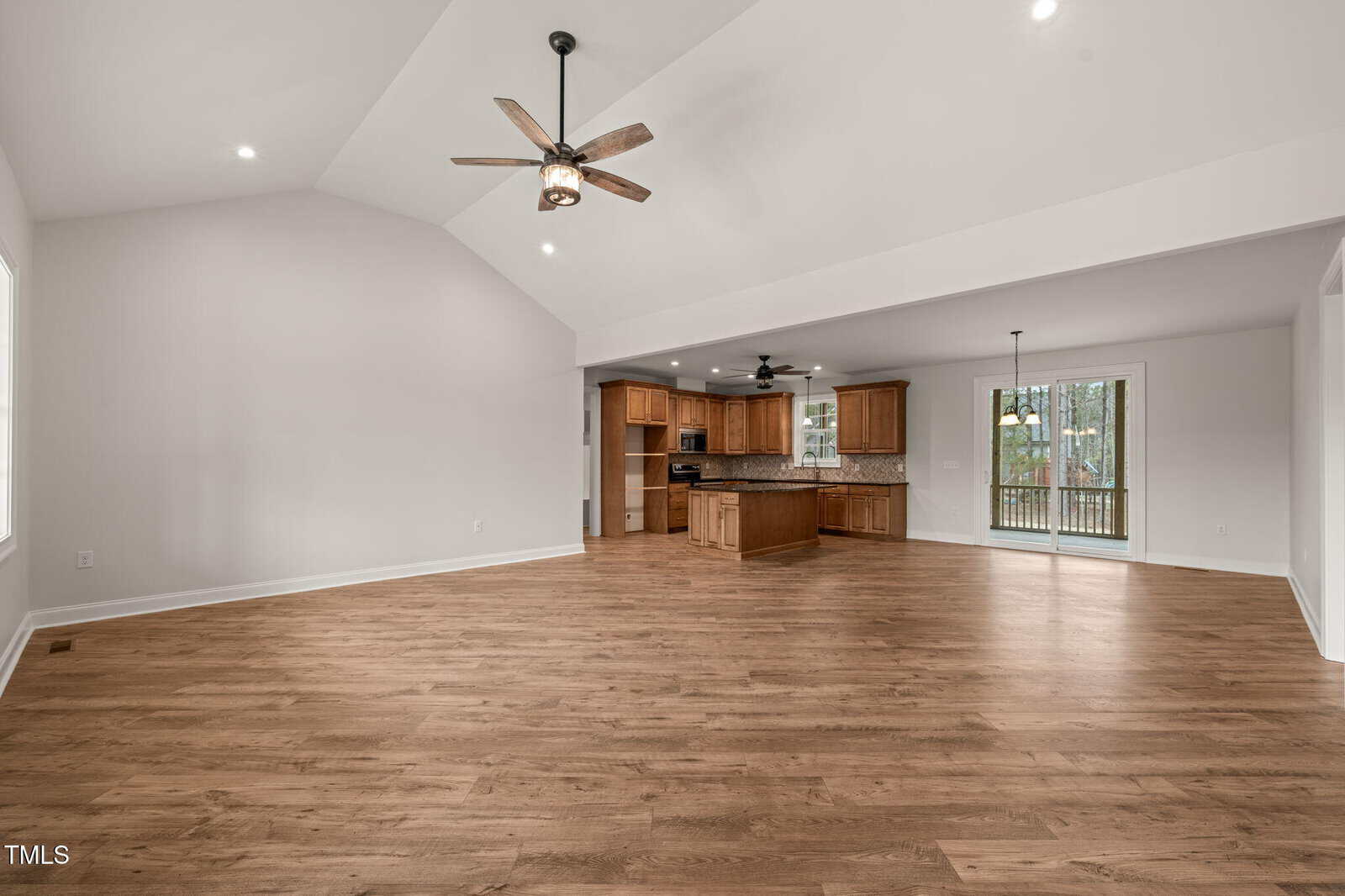 Spacious open-concept living area with light wood flooring, vaulted ceiling, ceiling fans, recessed lighting, and a kitchen with wooden cabinets and a center island in the background. Large windows provide natural light.