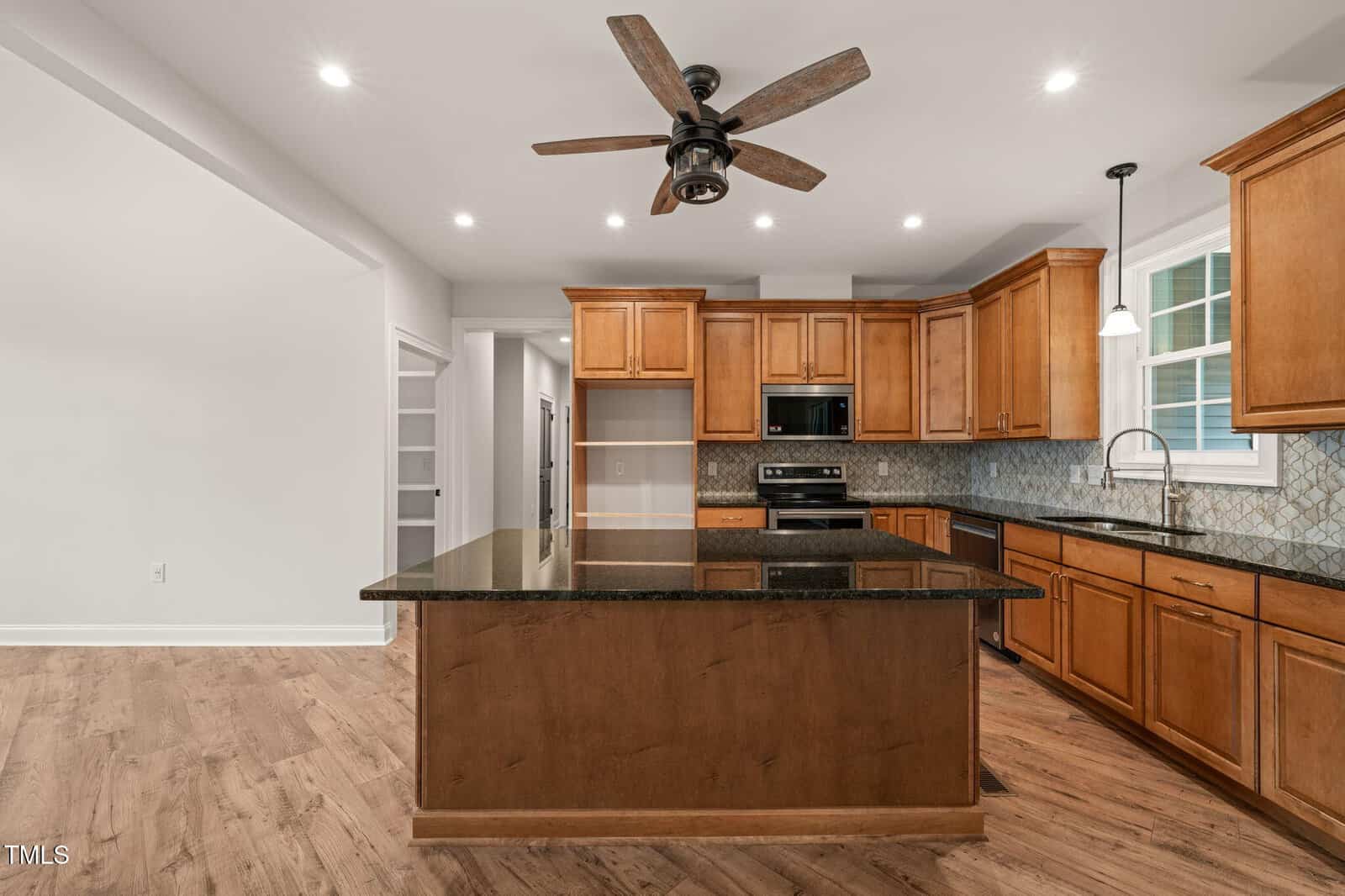 Spacious modern kitchen with wooden cabinets, a large central island with dark countertops, stainless steel appliances, tiled backsplash, wood flooring, ceiling fan, and recessed lighting.