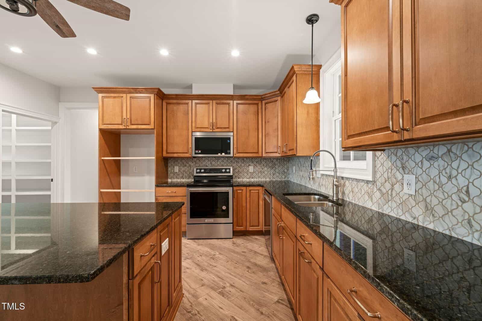 Modern kitchen featuring wood cabinets, black granite countertops, stainless steel appliances, a central island, and a patterned tile backsplash. A pendant light hangs over the sink next to a window.