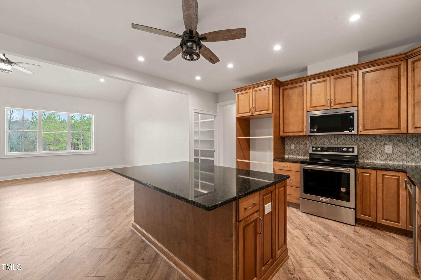 Modern kitchen with wood cabinets, black granite island, built-in appliances, wood flooring, ceiling fan, and adjacent open living area with large window and bright natural light.