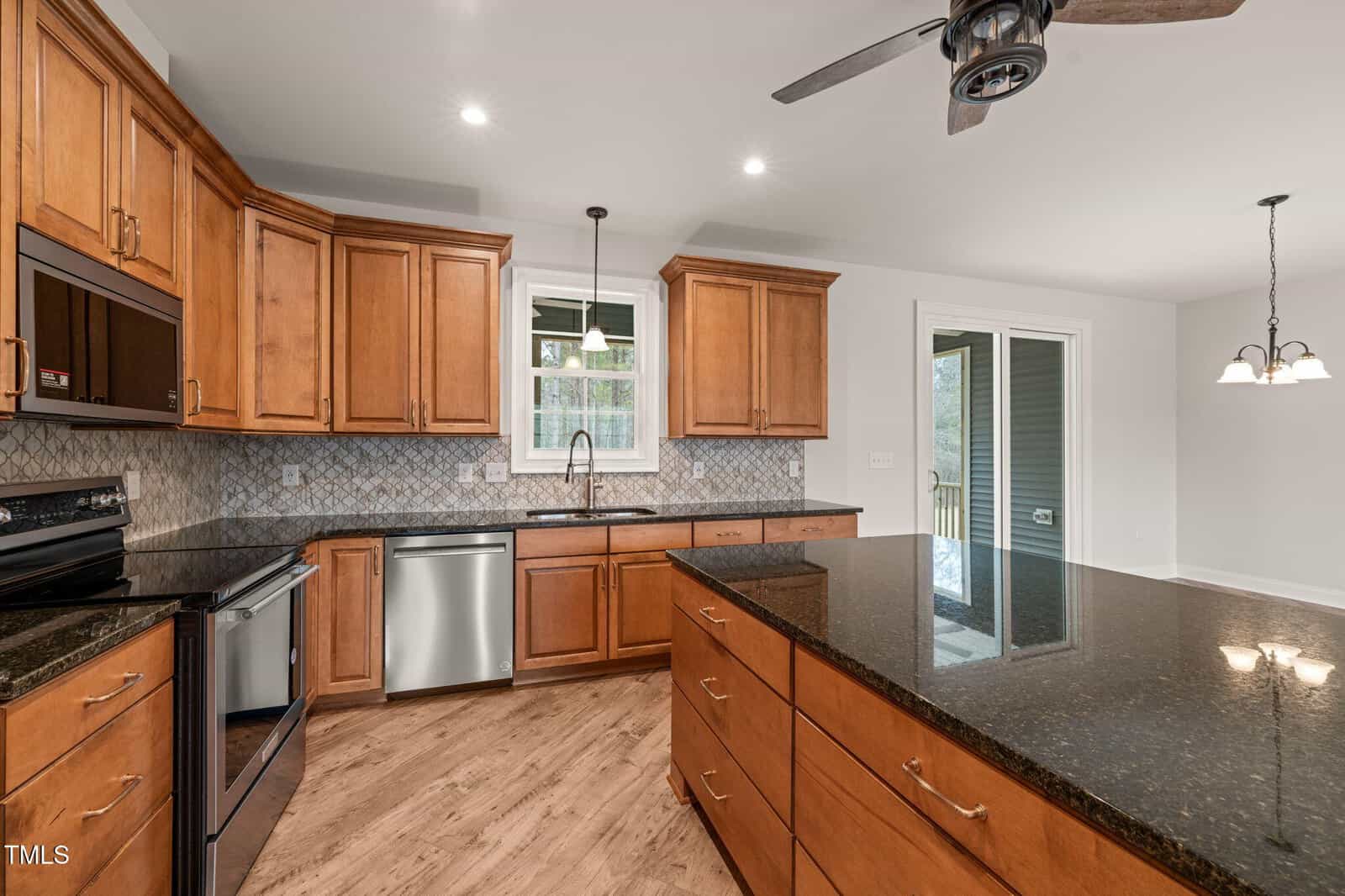 Spacious kitchen with wooden cabinets, granite island, stainless steel appliances, tile backsplash, and wood flooring. A window is above the sink, and sliding glass doors lead to an outdoor area. Ceiling fan and lights are visible.