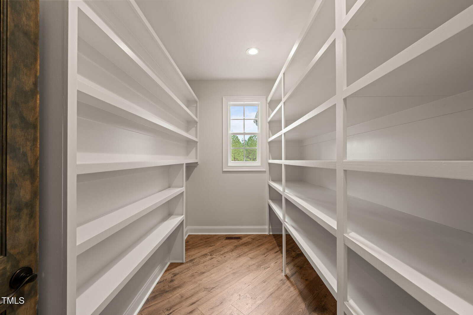 A spacious walk-in pantry with white built-in shelves on both sides, a wooden floor, and a small window letting in natural light at the end of the room.
