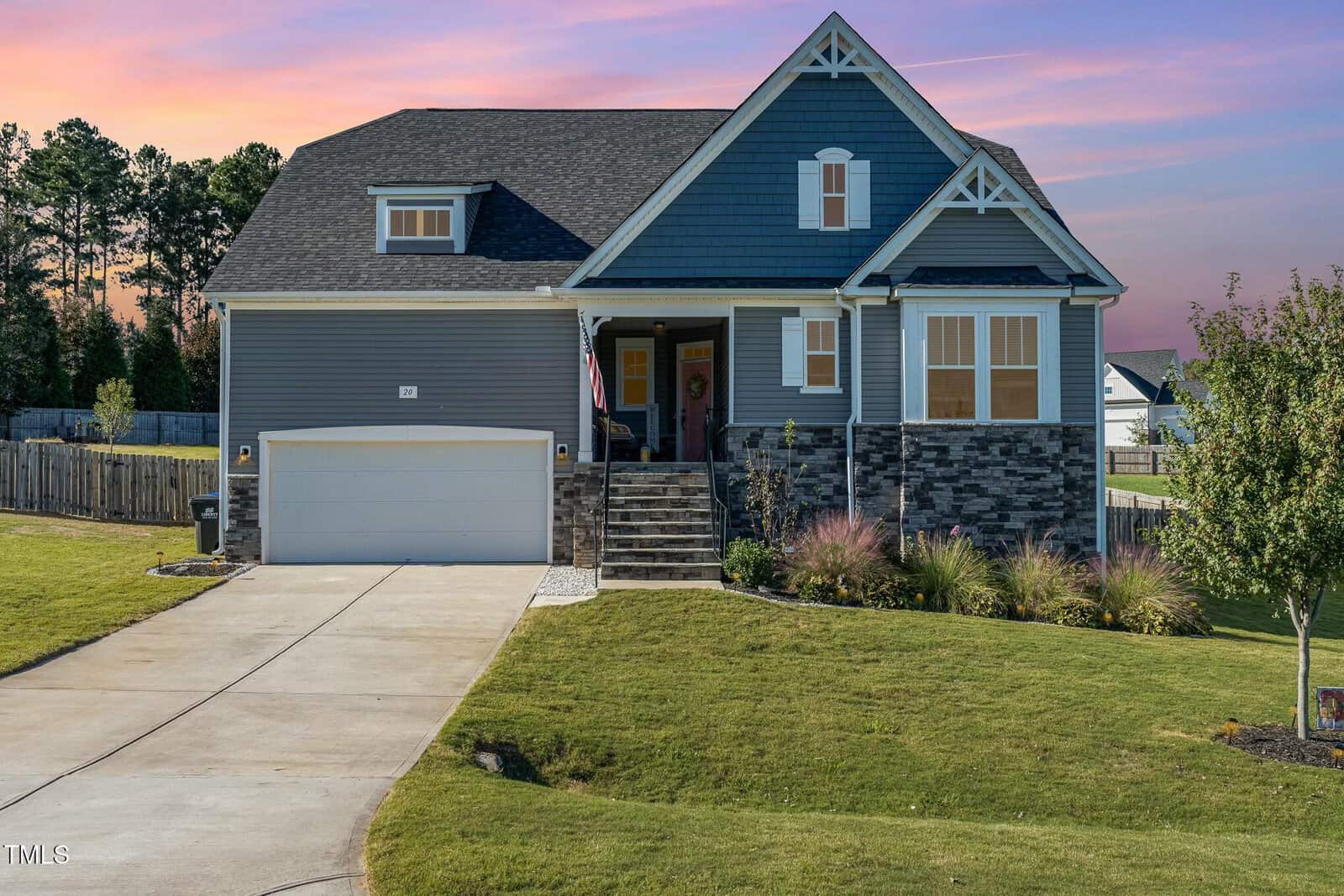 A two-story blue house with a stone accent, a front porch, and an attached two-car garage. The home sits on a grassy lawn with landscaping and a concrete driveway, with trees and a wooden fence in the background at sunset.
