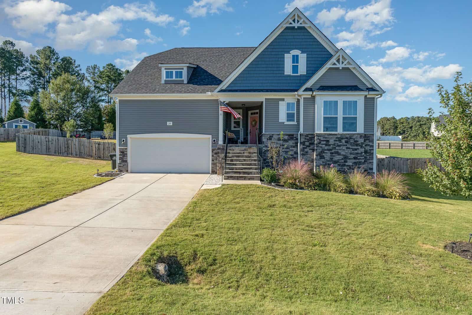 A two-story modern house with gray siding, a front porch, and an American flag. There is a double garage, a sloped driveway, a manicured lawn, and a fenced backyard. The sky is mostly clear with a few clouds.