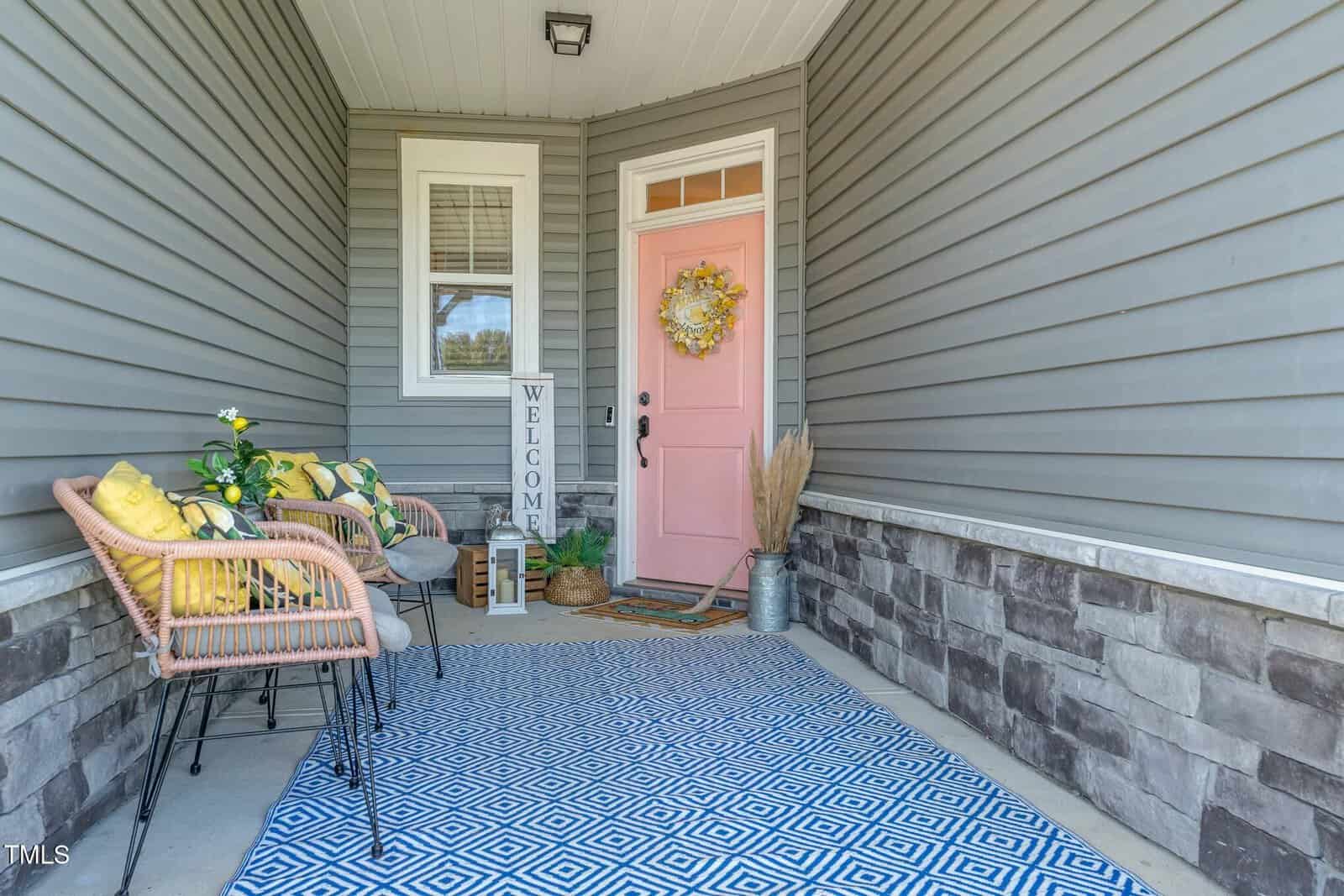 A covered porch with gray siding, a pink front door, and a blue patterned rug. Wicker chairs with pillows and plants sit beside a "WELCOME" sign. A wreath and doormat decorate the entry.