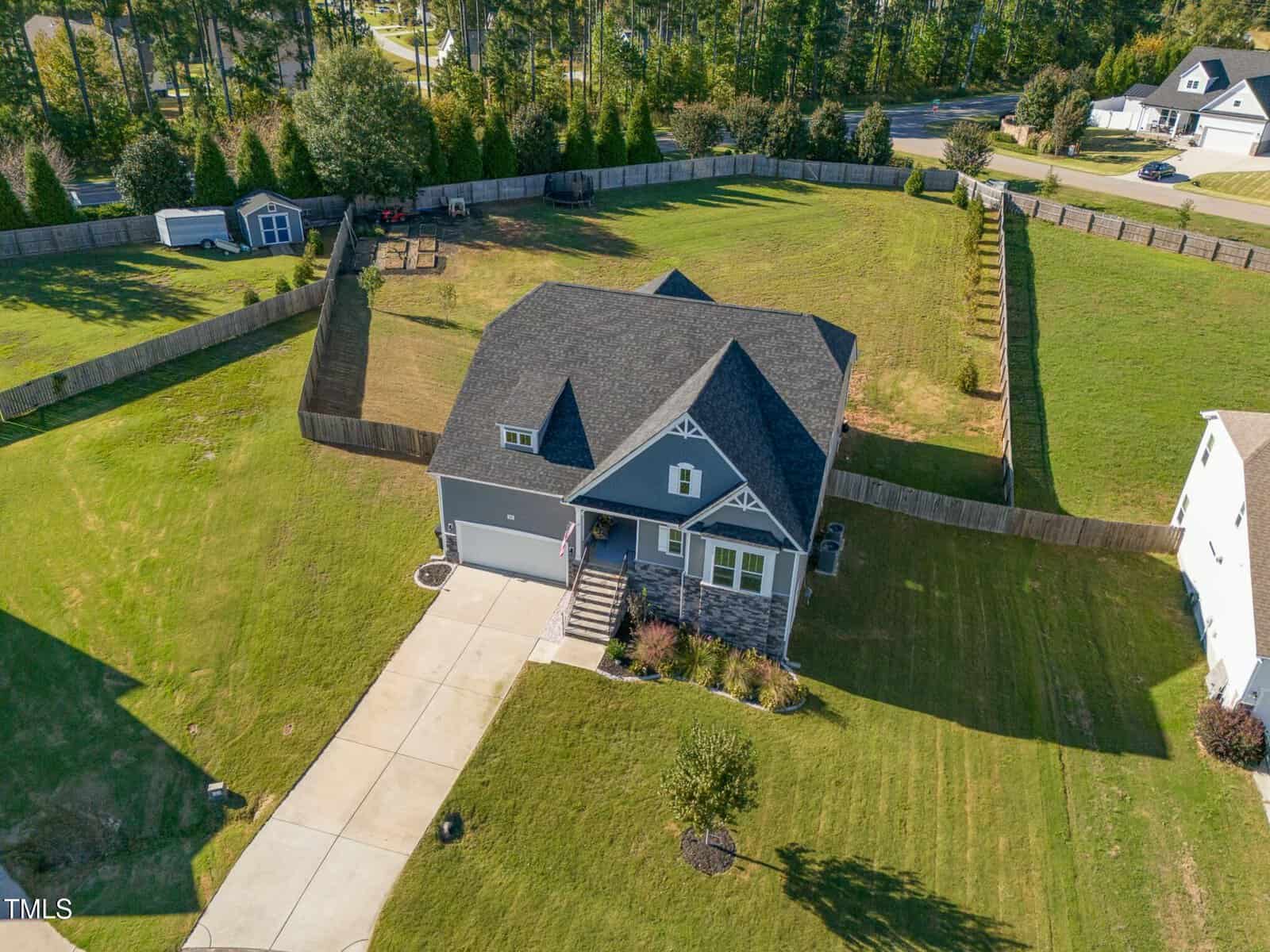 Aerial view of a single-family house with a gray roof, attached garage, and front porch, situated on a large, fenced, grassy lot with trees and a small shed in the backyard. Driveway leads to the street.