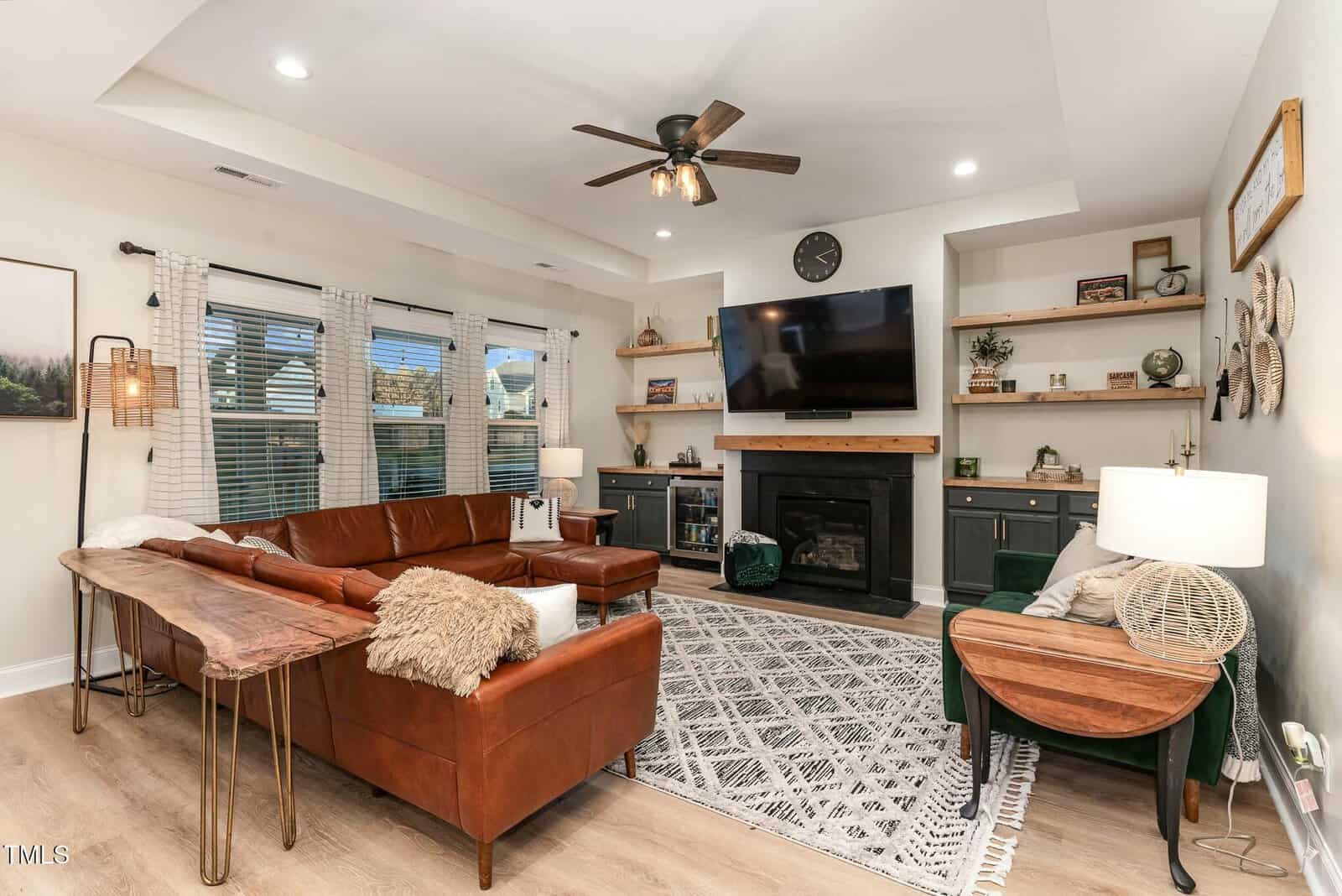 Modern living room with brown leather sofa, wooden tables, a fireplace under a wall-mounted TV, shelves with decor, large windows with curtains, and a patterned rug on wooden floors. Ceiling fan and recessed lighting above.