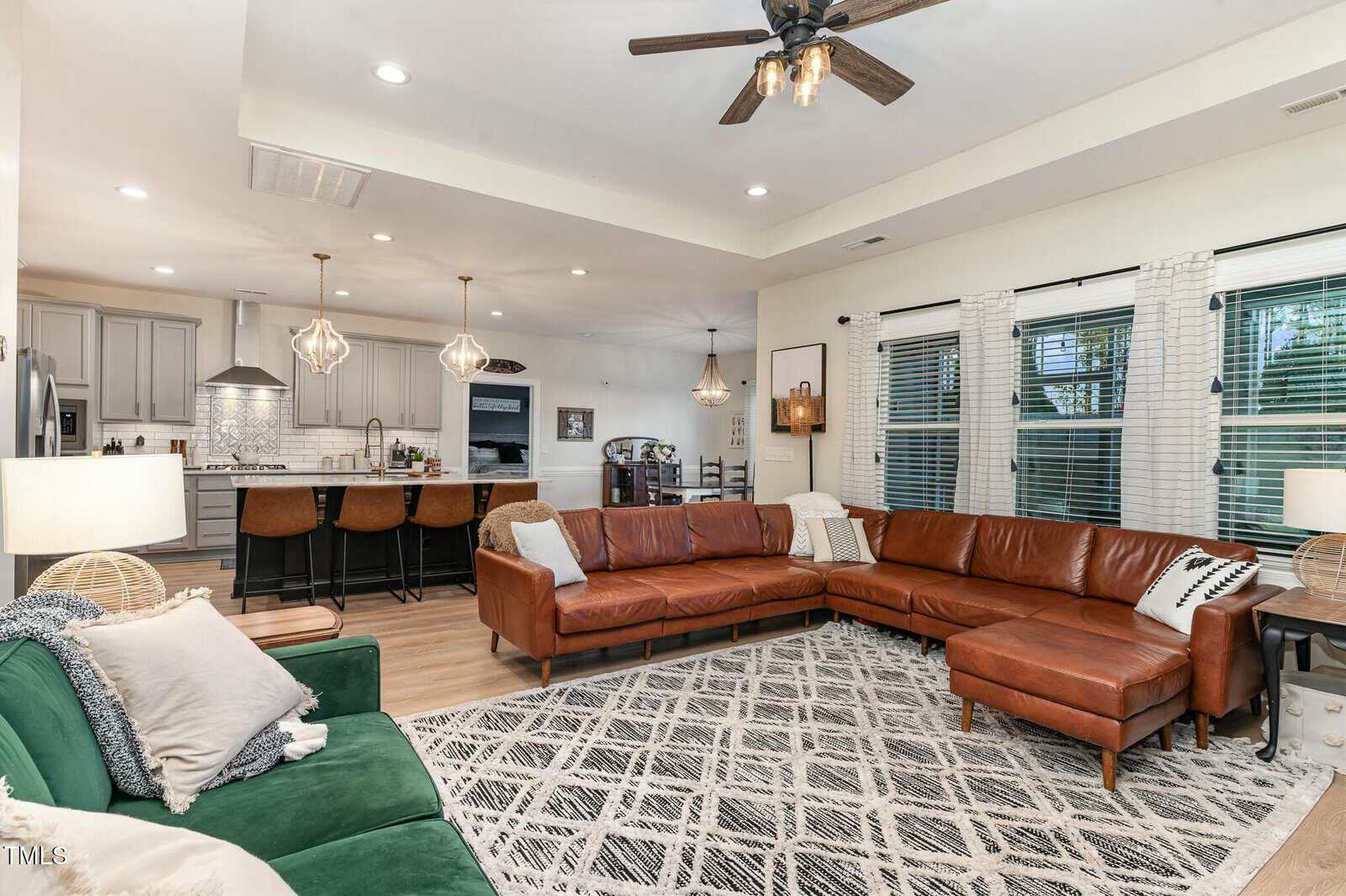 Modern open-concept living room with brown leather sectional, green sofa, patterned rug, and large windows. Kitchen with island and barstools in the background. Neutral tones and natural light fill the space.