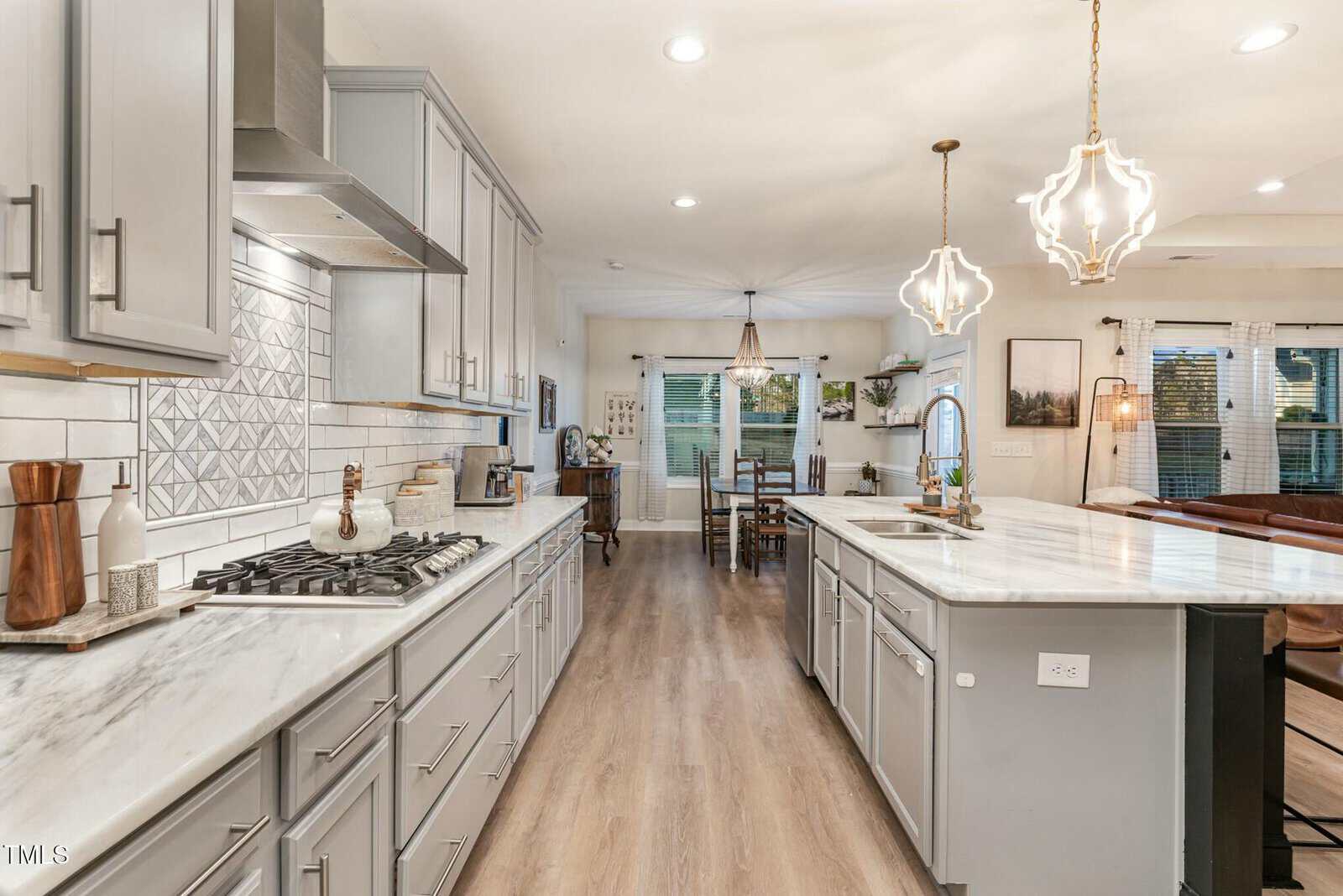 A modern kitchen with gray cabinets, stainless steel appliances, a marble island with pendant lights, and an open floor plan leading to a dining area with large windows.