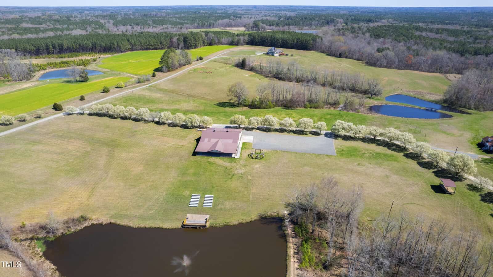 Aerial view of a rural landscape featuring a brown-roofed house surrounded by green fields and trees. A pond with a small dock is in the foreground, and another pond is visible to the right. Roads and fields stretch into the distance.