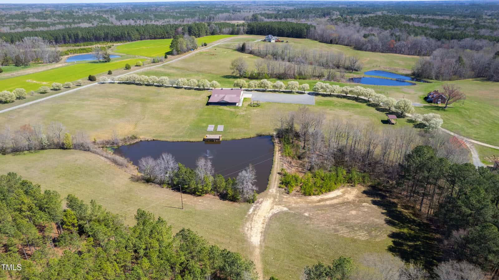 Aerial view of the rural landscape at 3200 Bliss Trail, featuring a large pond surrounded by trees, a red-roofed barn, and several small buildings. Open fields and scattered trees stretch into the distance under a blue sky, embodying the tranquility of this scenic stem of nature.