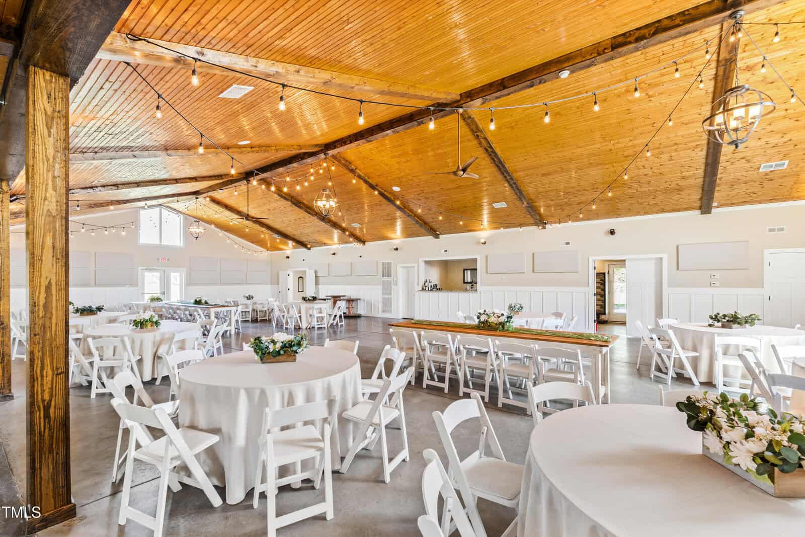 Spacious event hall with a wooden ceiling and string lights. Round tables covered with white tablecloths and white chairs are arranged throughout. There are floral centerpieces on the tables, and a long wooden table is set up against the wall.