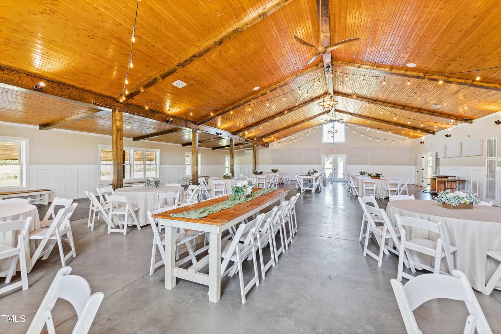 A large, well-lit event hall with a wooden ceiling and floor. The room is set up with round and rectangular tables covered in white tablecloths, surrounded by white chairs. String lights and a chandelier hang from the ceiling, and floral centerpieces adorn the tables.