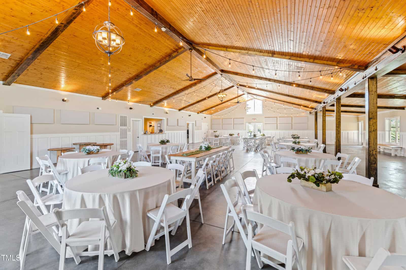 A spacious event hall with a wooden ceiling and string lights features round tables covered with white tablecloths and arranged with floral centerpieces. White chairs surround the tables, and there are large windows along the walls.