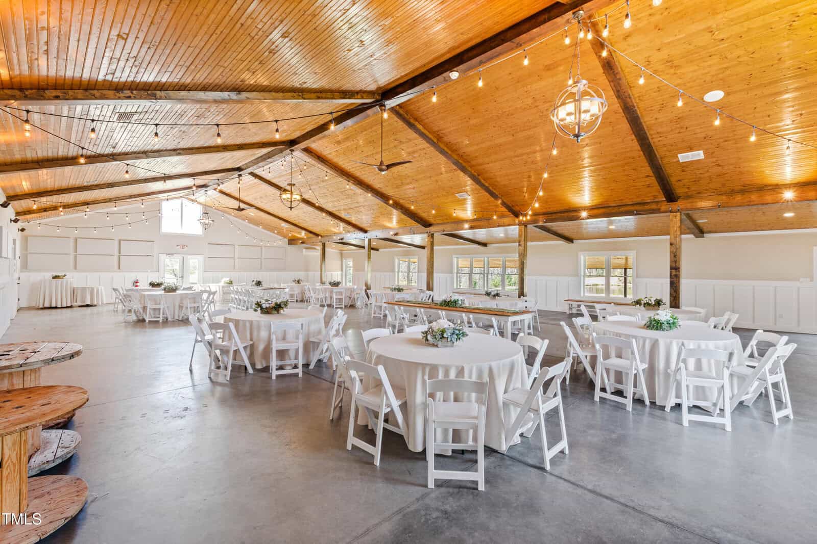 A spacious event hall with a wooden ceiling and exposed beams. Round tables covered with white tablecloths are set up with chairs around them. String lights hang overhead. Small floral centerpieces decorate the tables. Large windows let in natural light.