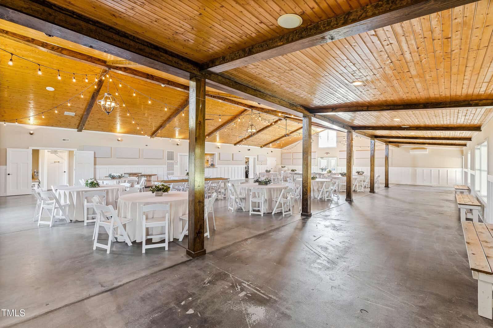 A spacious indoor event venue with a wooden ceiling and concrete floor. Round tables covered with white tablecloths and white chairs are arranged throughout. String lights hang overhead, and natural light fills the room from large windows.
