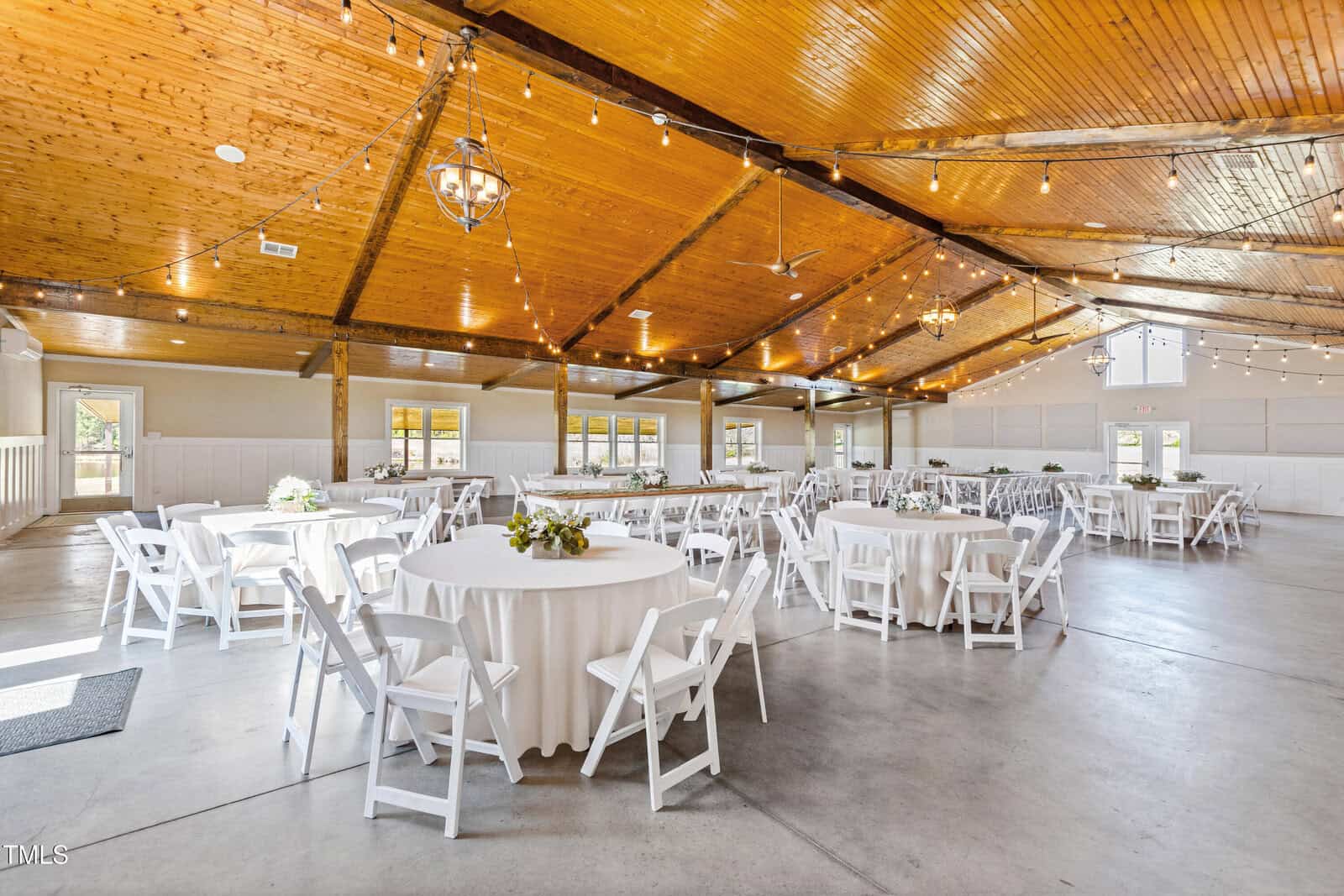 A spacious event venue with a wooden ceiling and string lights. Several round tables are draped with white tablecloths, each surrounded by white chairs. Floral centerpieces are placed on the tables. Large windows allow natural light to fill the room.
