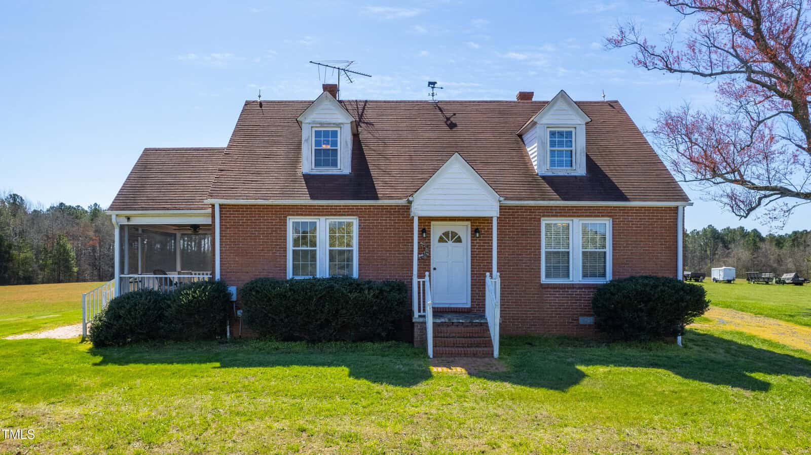A red brick house with a gabled roof and two dormer windows sits on a grassy lawn. The front features a small porch with white railings. A large tree is partially visible on the right, and the background shows a clear sky and wooded area.