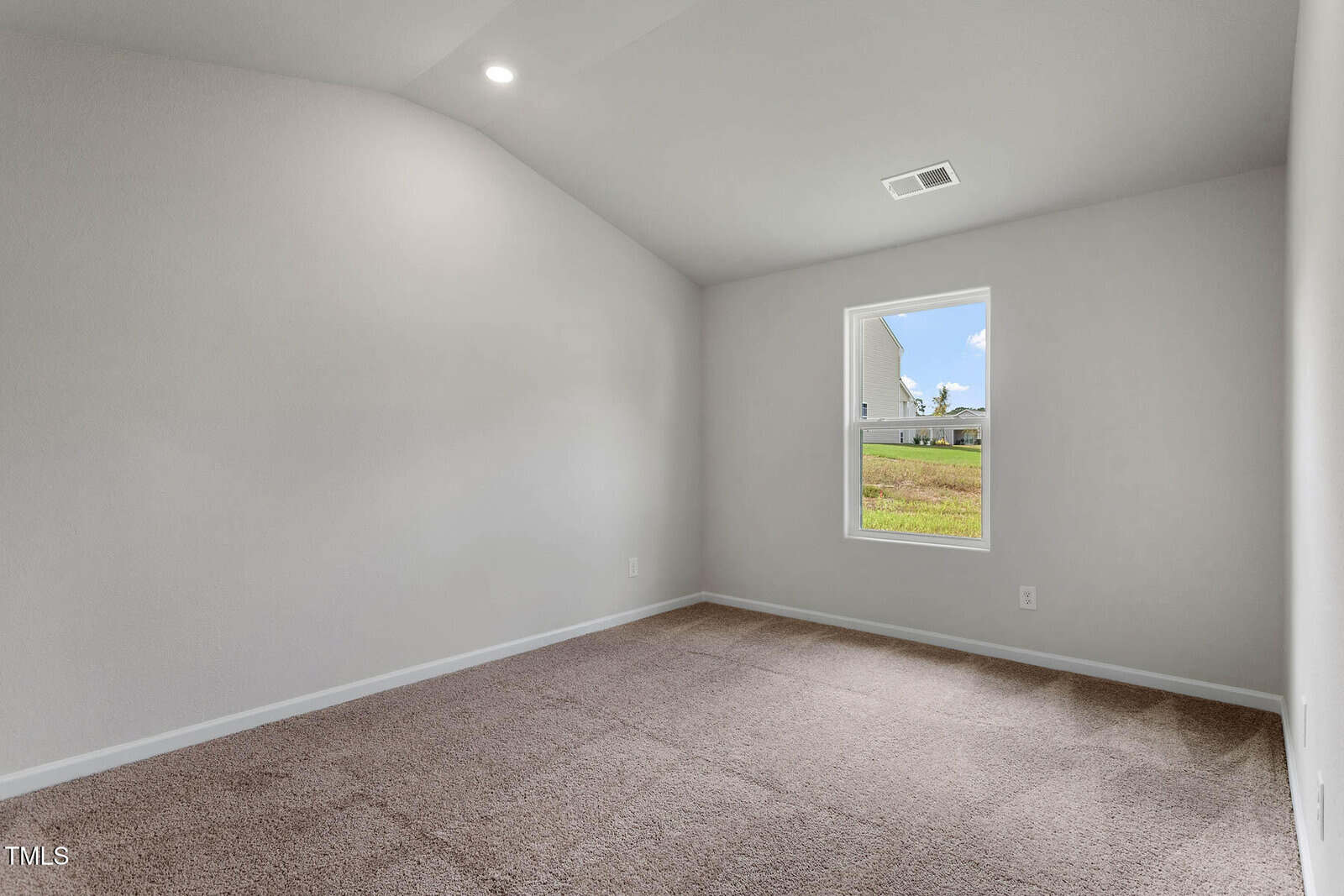 An empty room with carpeted flooring and white walls features a single window showing a view of an exterior building and grass. The ceiling is slightly vaulted, and the space is illuminated by a ceiling light.