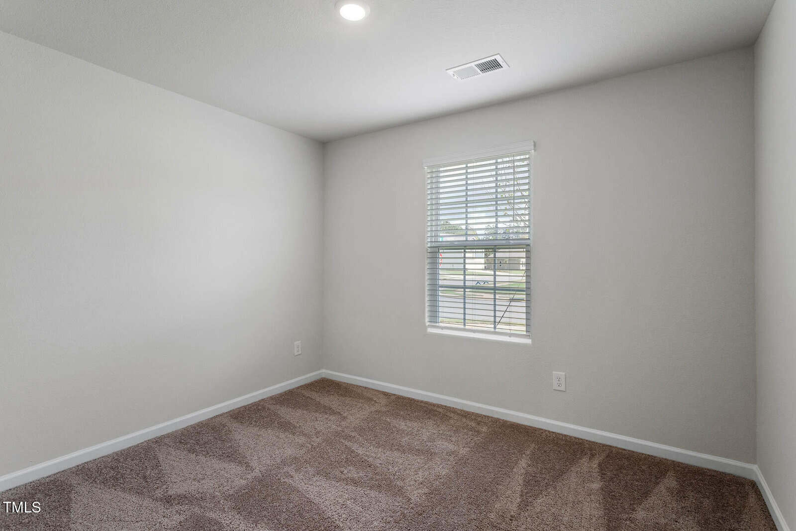 An empty room with beige walls and a beige carpeted floor. A single window with blinds lets in natural light. There's a ceiling light fixture, and a power outlet is visible on the wall.