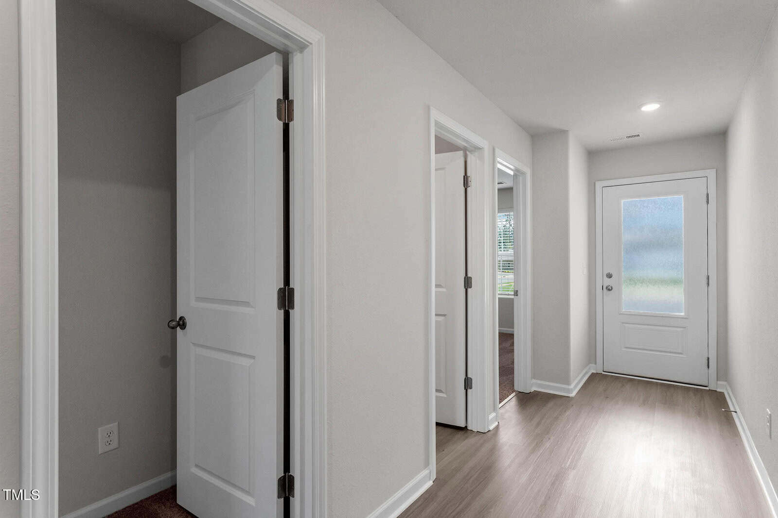 A hallway with light-colored walls and wood flooring, featuring three white doors on the left side and a glass-paneled door at the end. Natural light enters through the doors, illuminating the space.