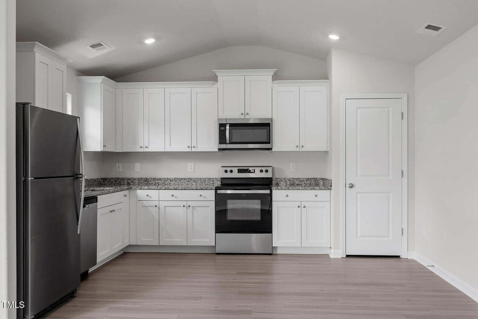 A modern kitchen with white cabinets and stainless steel appliances, including a refrigerator, stove, and microwave. The countertop is granite, and the flooring is a light wood. There is a closed white door to the right. Lighting is recessed.