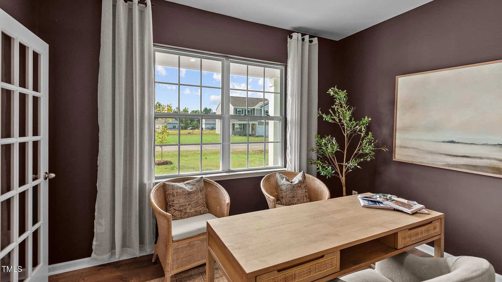 A home office with dark brown walls, a wooden desk, two wicker chairs with cushions, a potted plant, a large window with white curtains, a landscape painting, and a stack of magazines on the desk.